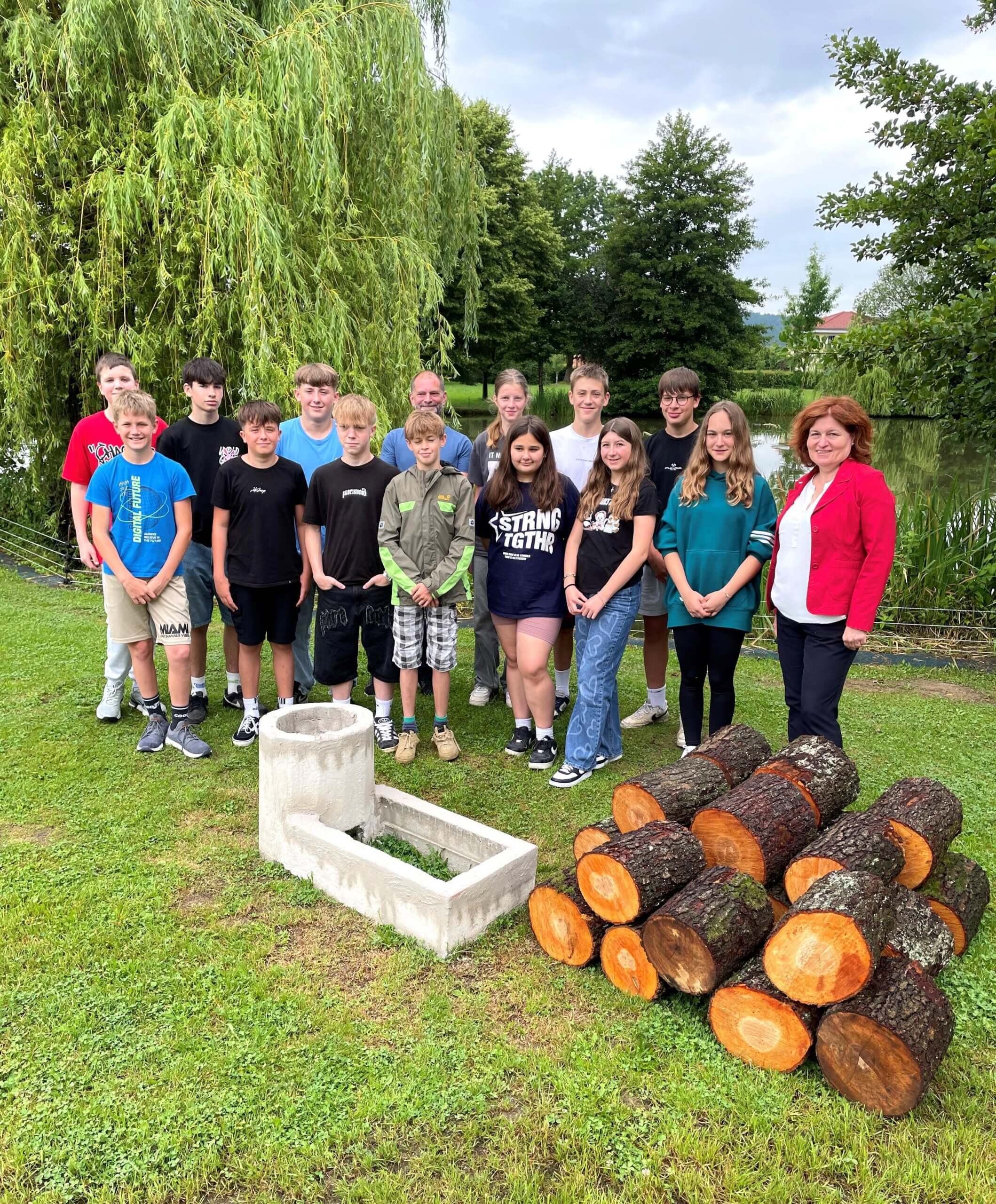 Gruppe von Jugendlichen und Erwachsenen posiert im Freien neben Holzstämmen und Steinstruktur unter einem Baum.
