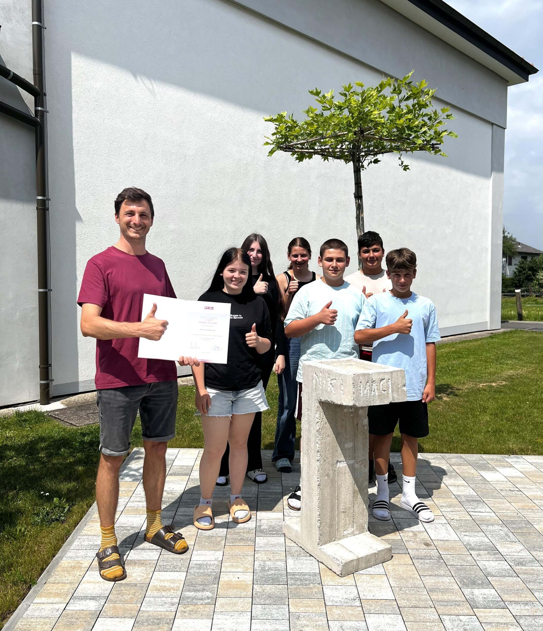 Lehrer und Schüler feiern eine Auszeichnung im Freien, Daumen hoch vor einem Beton-Aufsteller.
