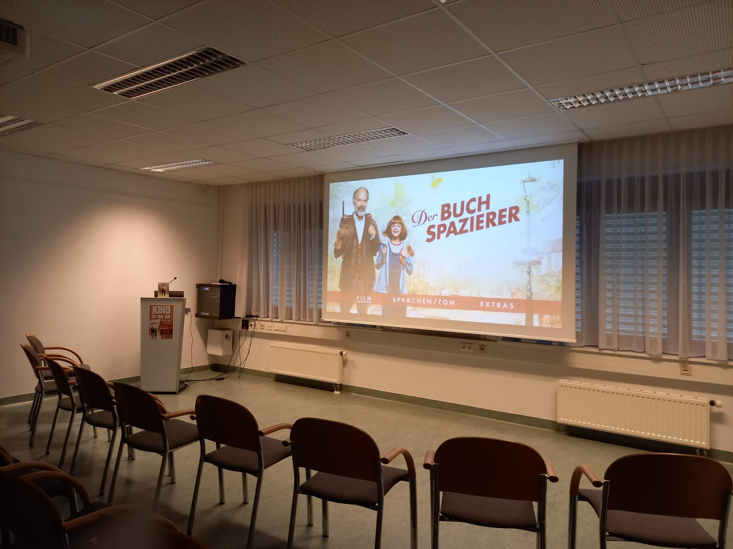 Leerer Seminarraum mit Filmplakat Der Buchspazierer auf Leinwand, Stühle und Rednerpult im Vordergrund.