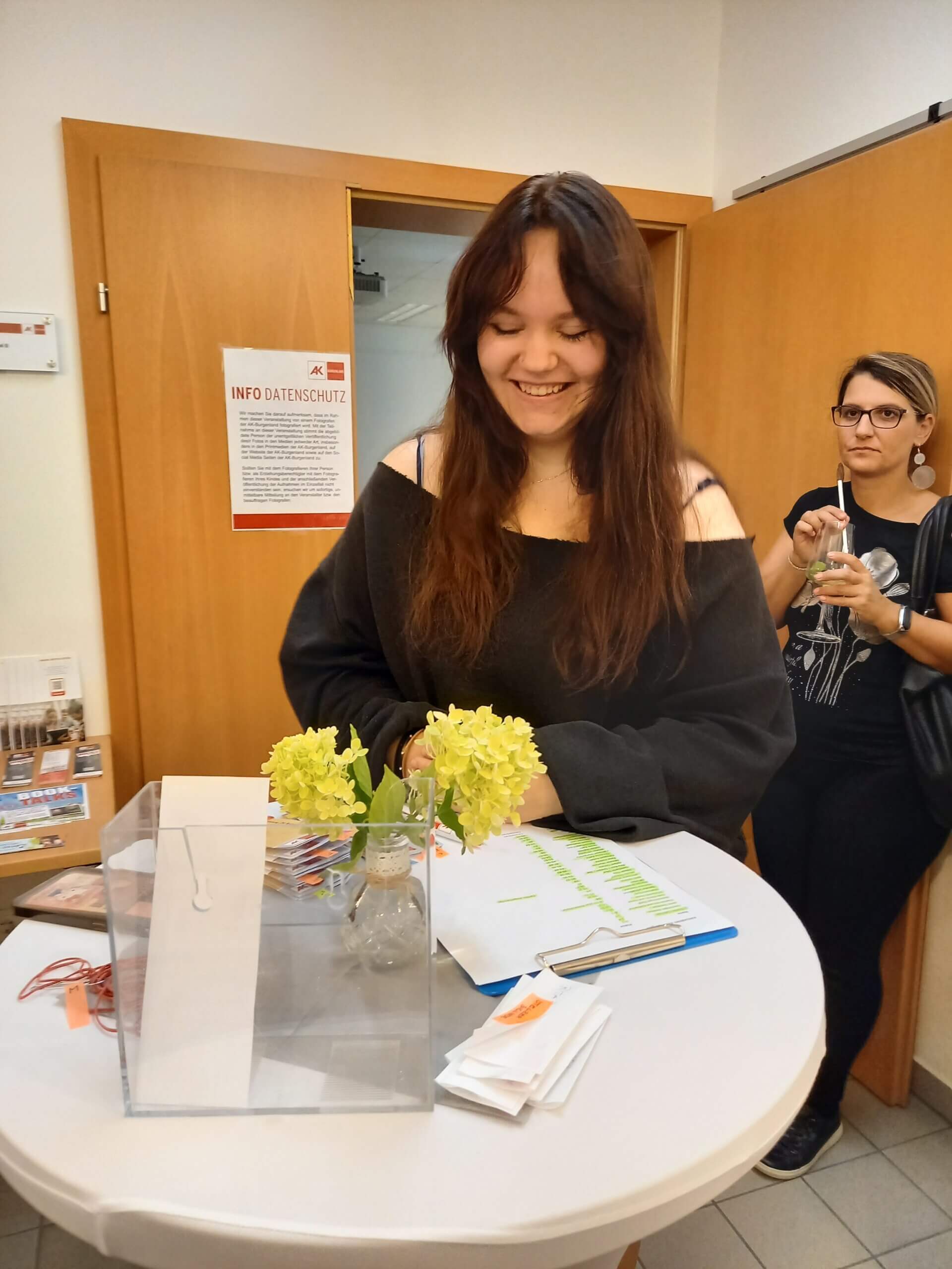 Eine lachende Frau steht an einem Tisch mit Blumen und Zettel in einem Büro oder Empfangsbereich.