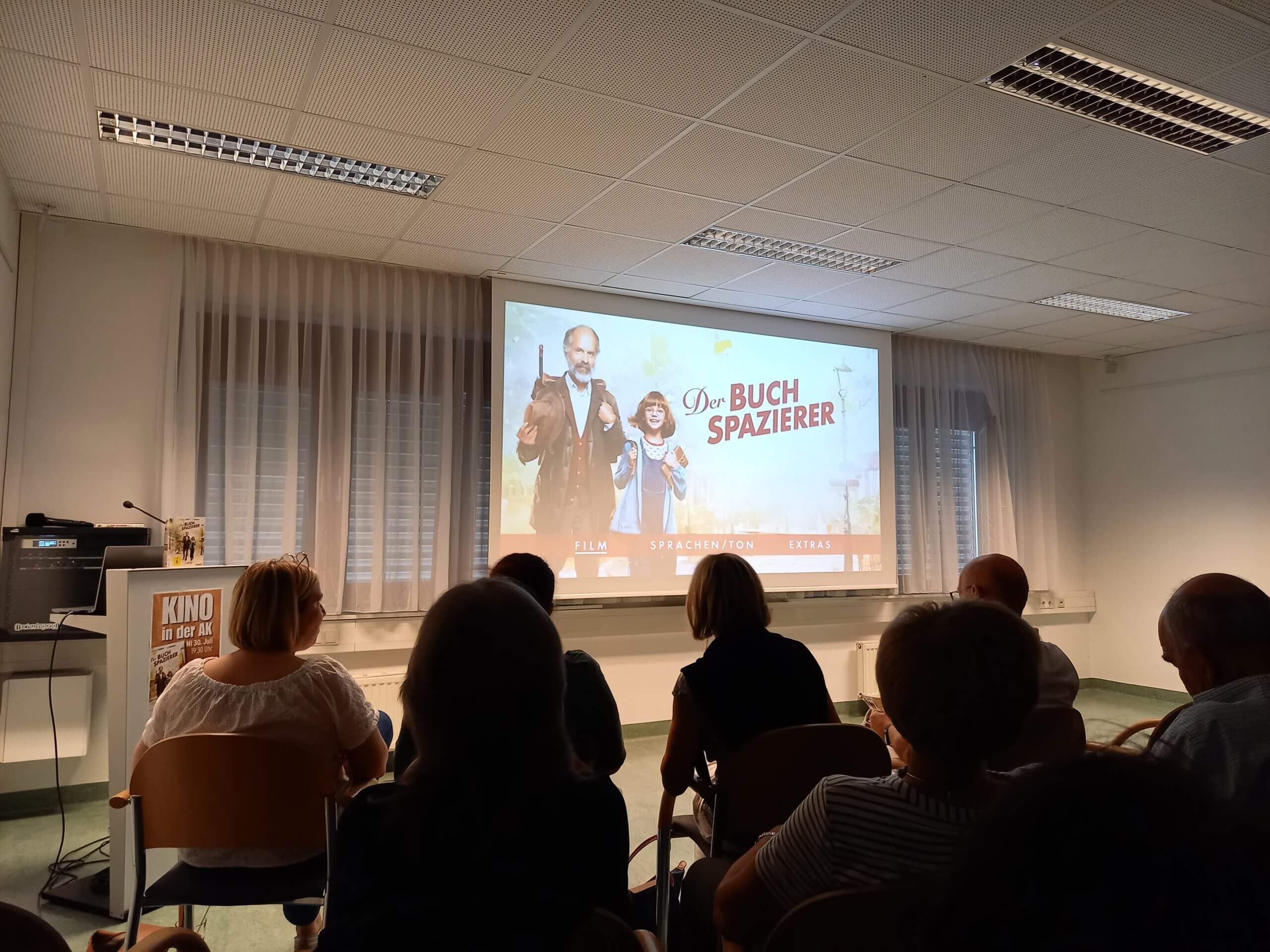 Leute schauen Der Buchspazierer im Kino; Bild von Leinwand mit Filmplakat, Publikum im Vordergrund.