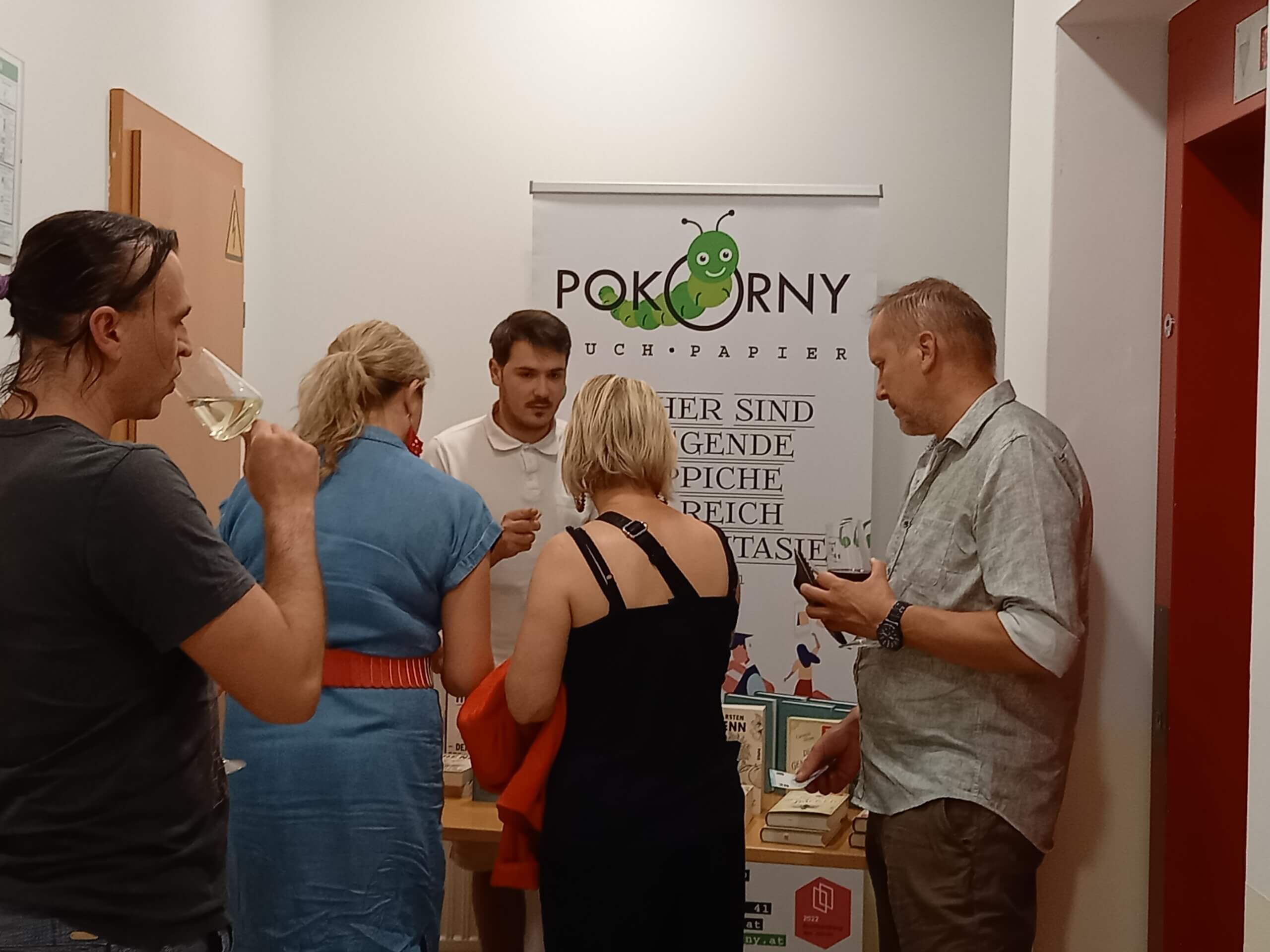 Menschen auf einer Buchvorstellung bei Pokorny Buch-Papier, diskutierend und Wein trinkend, vor einem Werbebanner.