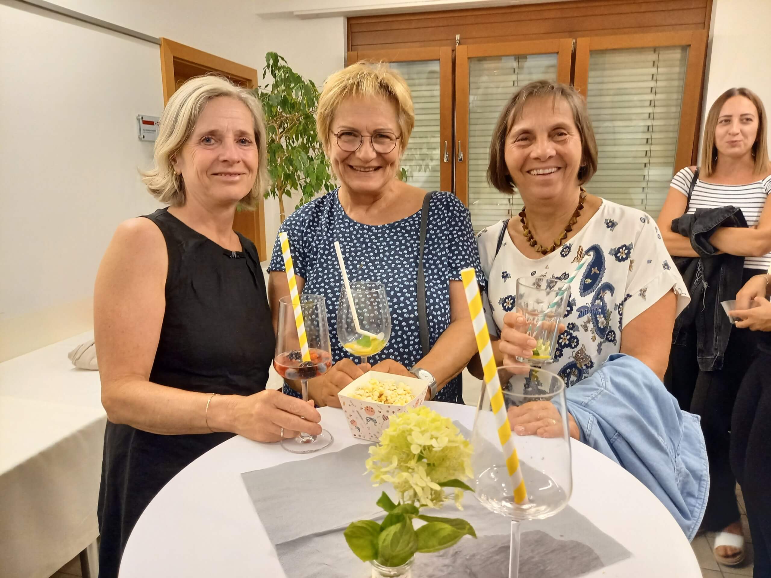 Drei Frauen bei einer Feier, lächelnd mit Getränken in der Hand, an einem Stehtisch mit Blumen.