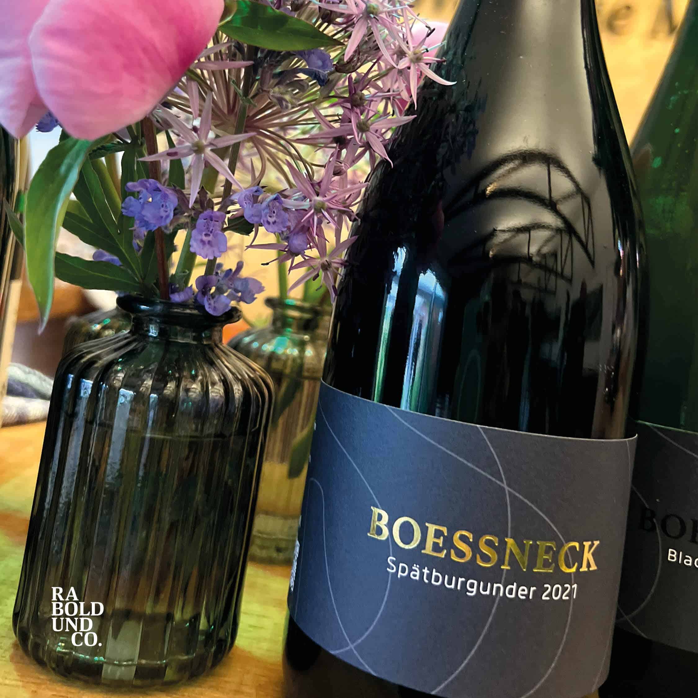 Flasche Boessneck Spätburgunder 2021 neben Blumenarrangement in Glasvase auf einem Tisch.