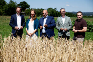 Eine Gruppe von Personen steht in einem Weizenfeld, trägt formelle Kleidung und lächelt bei sonnigem Wetter.