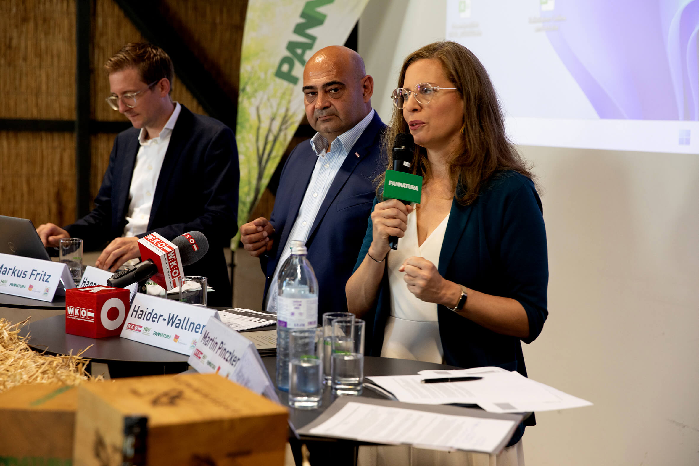 Drei Personen bei einer Pressekonferenz mit Mikrofonen von WKO und PANATURA, sprechen über ein wichtiges Thema.