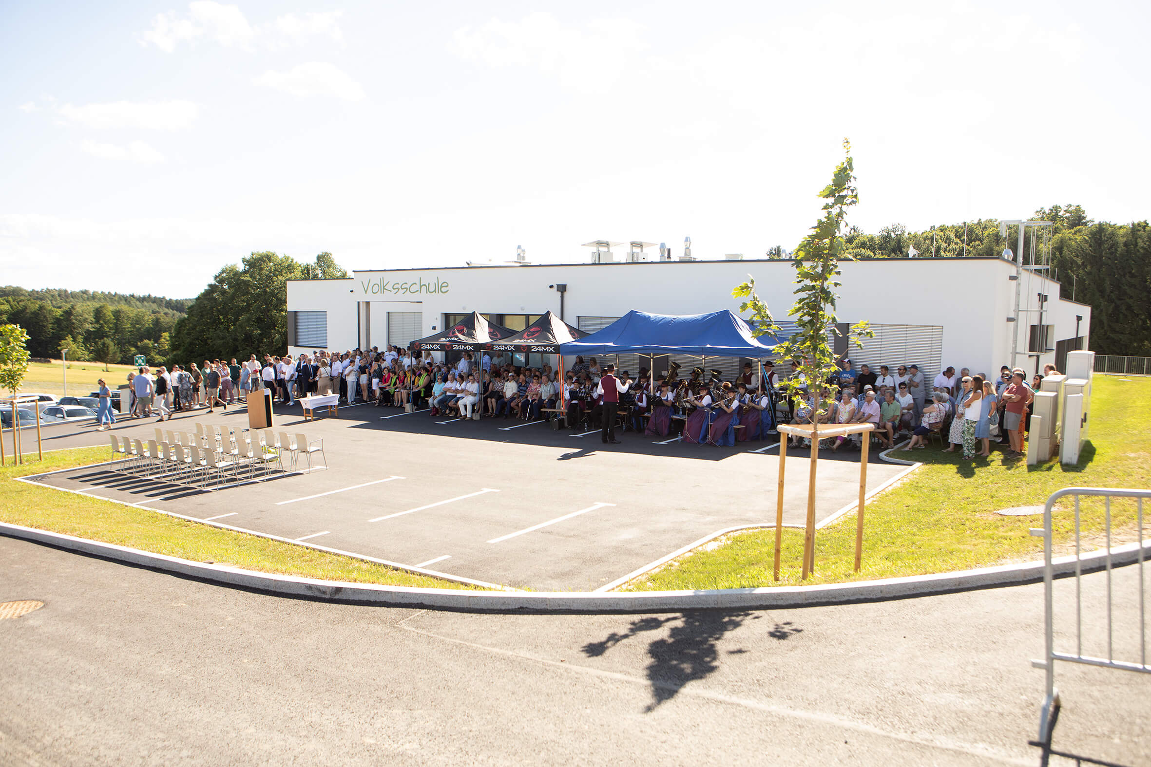 Versammlung vor einer Volksschule mit Zelt und Publikum bei einer Feier im Freien an einem sonnigen Tag.