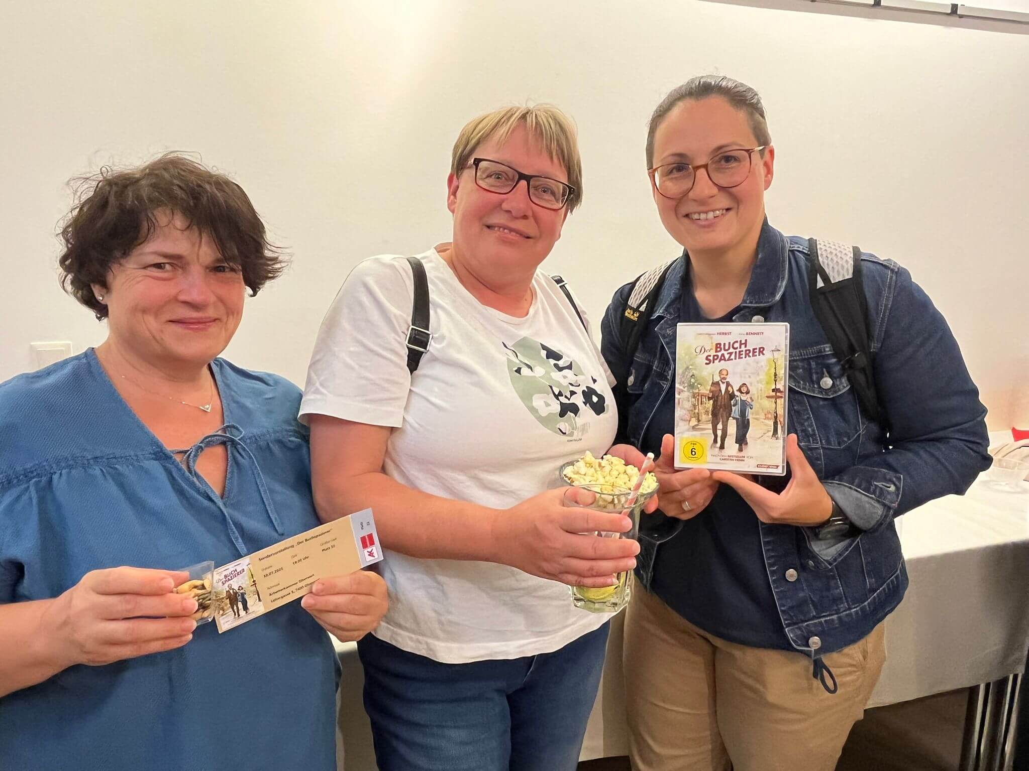 Drei Frauen halten ein Ticket, Popcorn und eine DVD von Der Buchspazierer und lächeln in die Kamera.