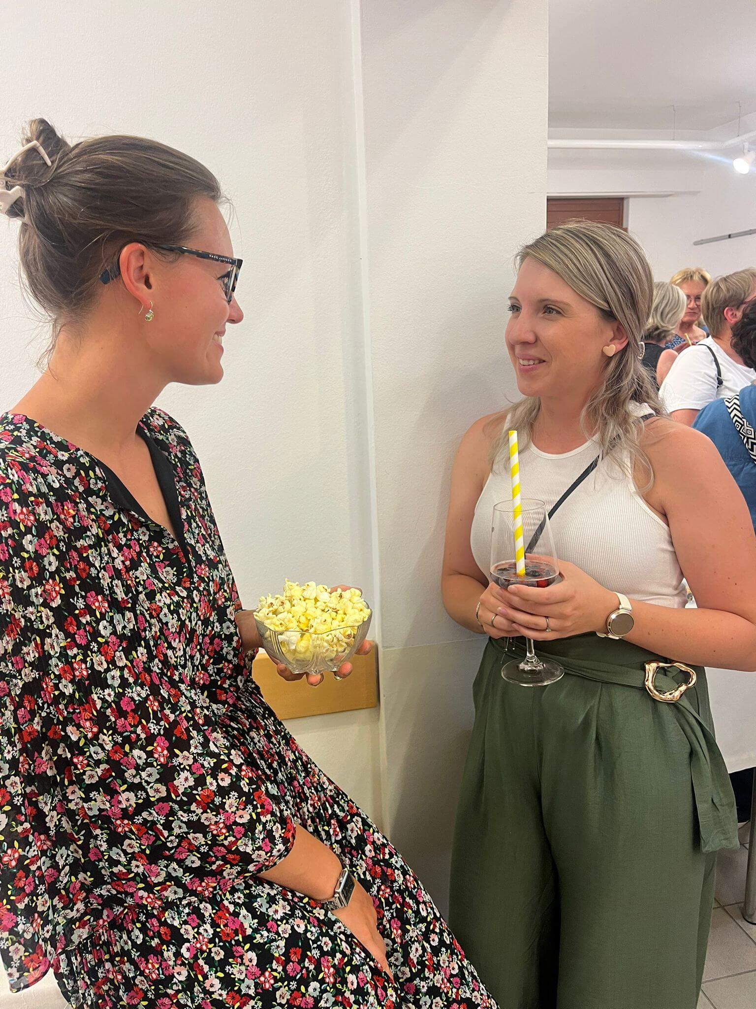 Zwei Frauen unterhalten sich bei einer Party mit Popcorn und Getränken, lächelnd in lockerer Atmosphäre.