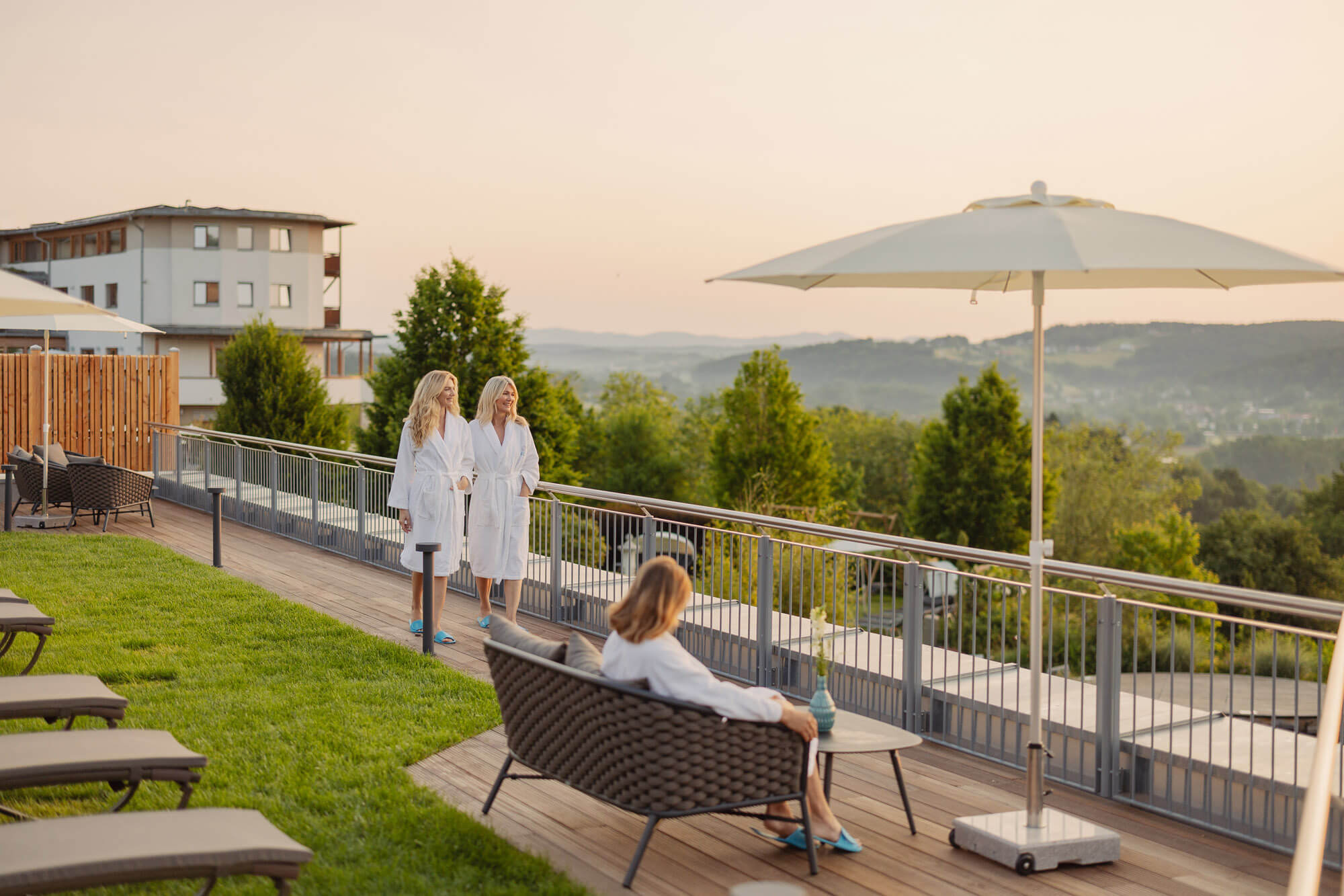 Frauen in Bademänteln genießen die Aussicht auf grüne Hügel von einer Terrasse aus bei Sonnenuntergang, entspannend im Spa.