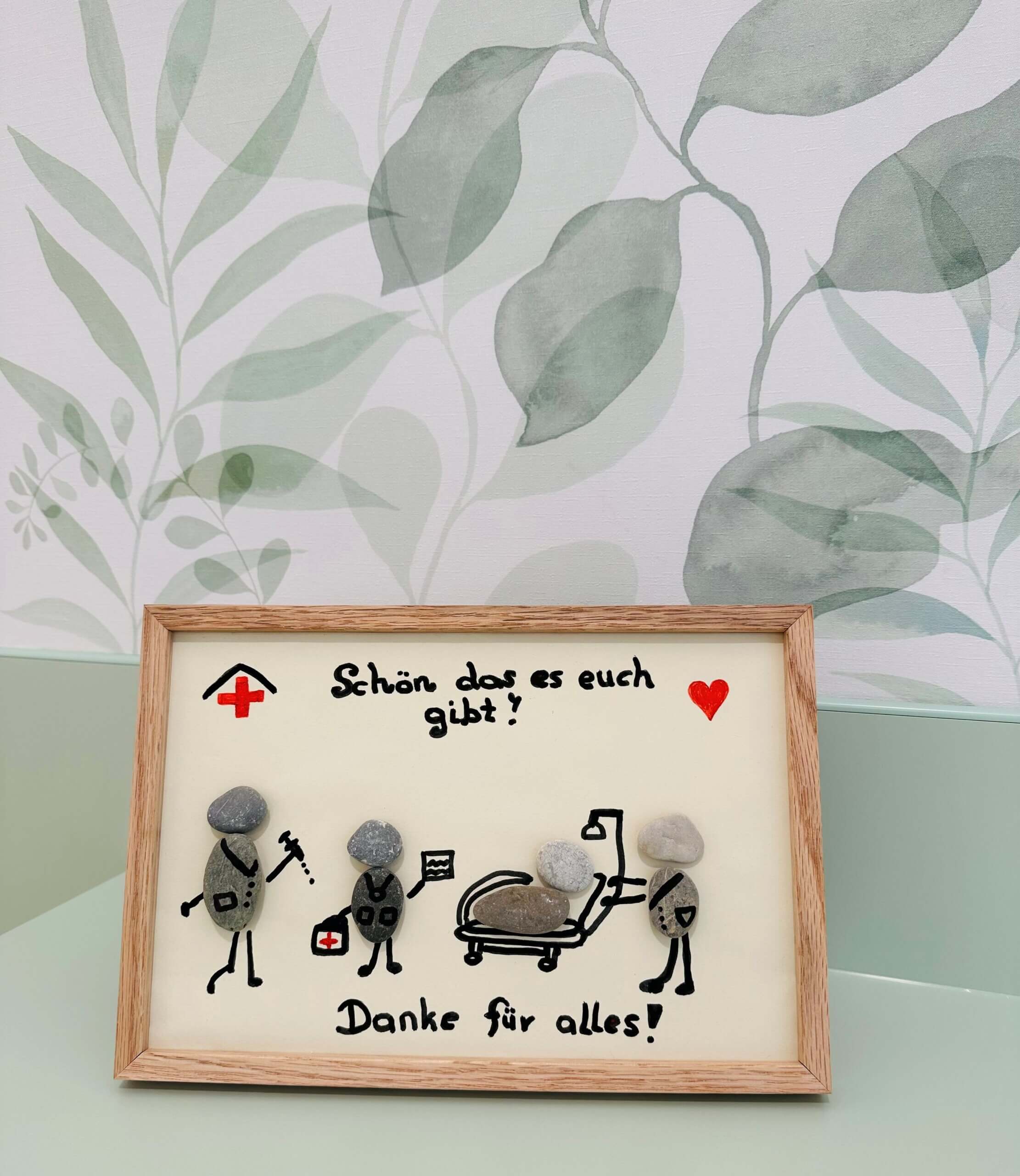 Danksagung an medizinisches Personal: Bild mit bemalten Steinen und Botschaft zur Wertschätzung, Holzrahmen mit Blattmotiv.