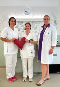 Drei medizinische Fachkräfte in weißer Kleidung halten ein rotes Herz in einer Klinikumgebung.