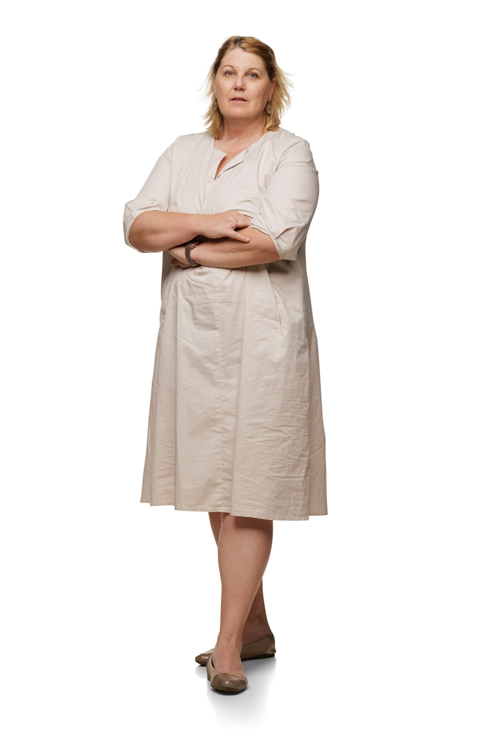 Frau in beigem Kleid steht mit verschränkten Armen vor weißem Hintergrund, neutraler Gesichtsausdruck.