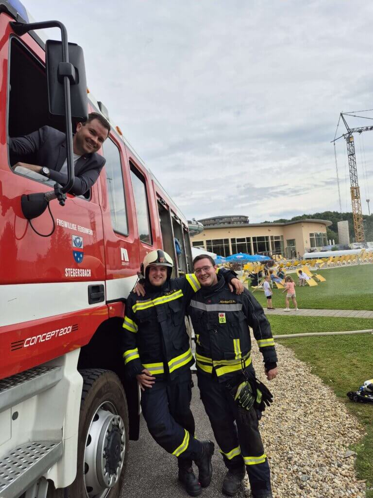 Feuerwehrleute in Einsatzkleidung neben einem roten Feuerwehrauto, fröhliche Szene im Freien mit Kindern im Hintergrund.