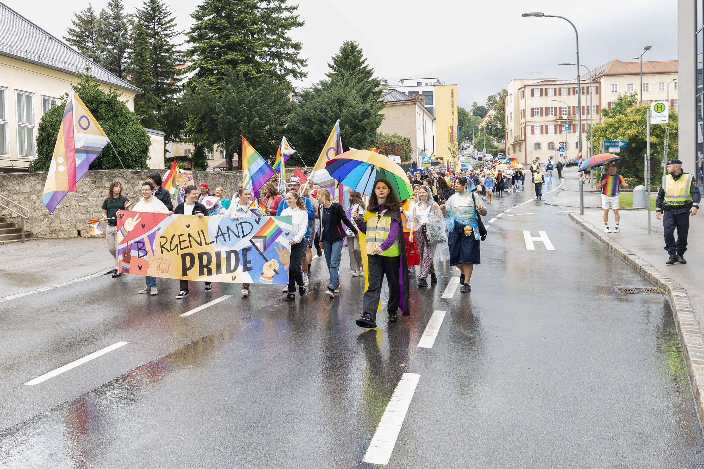 Teilnehmer*innen der Burgenland Pride Parade mit Regenbogenfahnen auf nasser Straße.