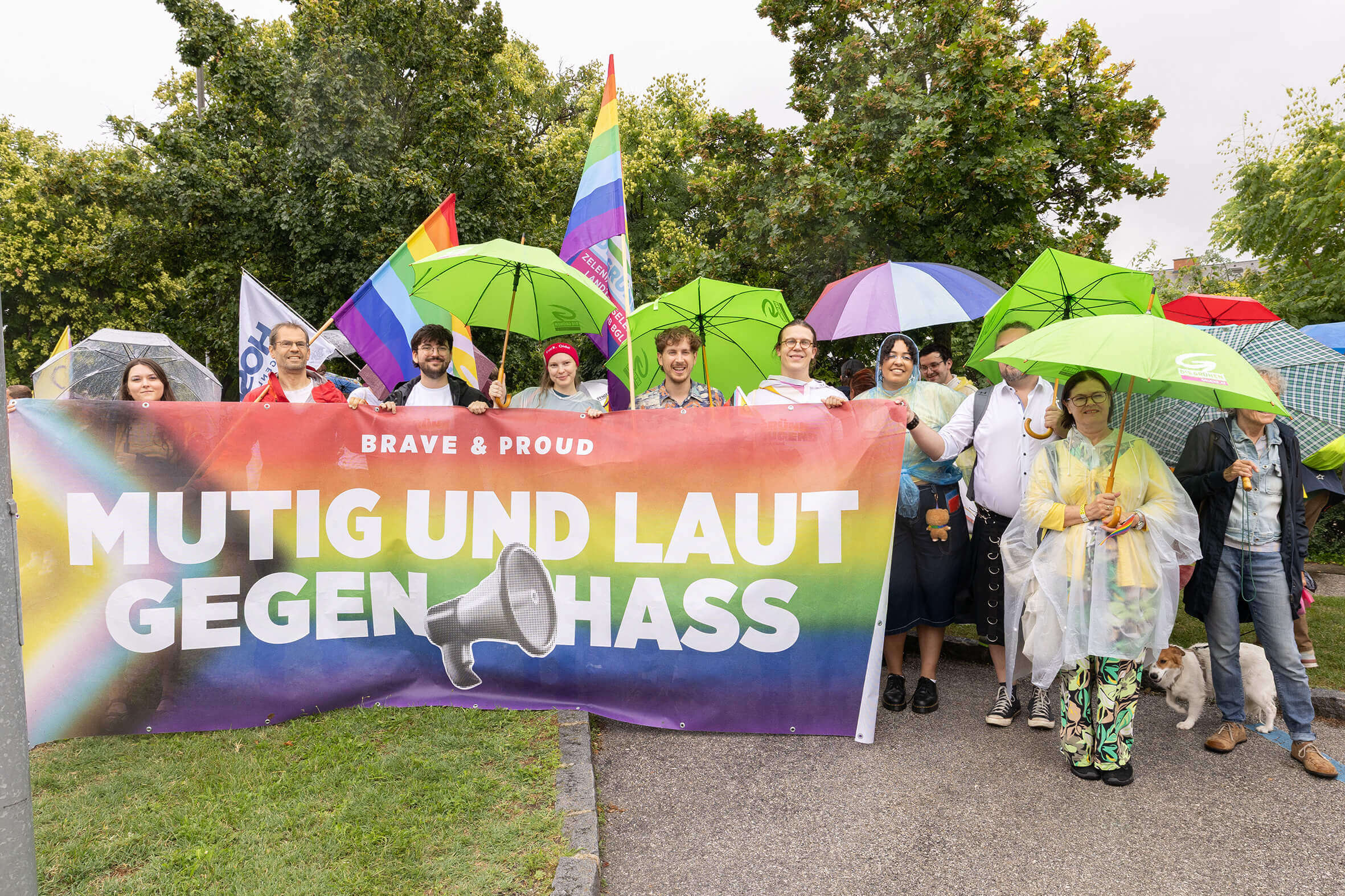 Gruppe bei Regen mit Mutig und laut gegen Hass-Banner, bunte Regenschirme und Regenbogenfahnen im Hintergrund.