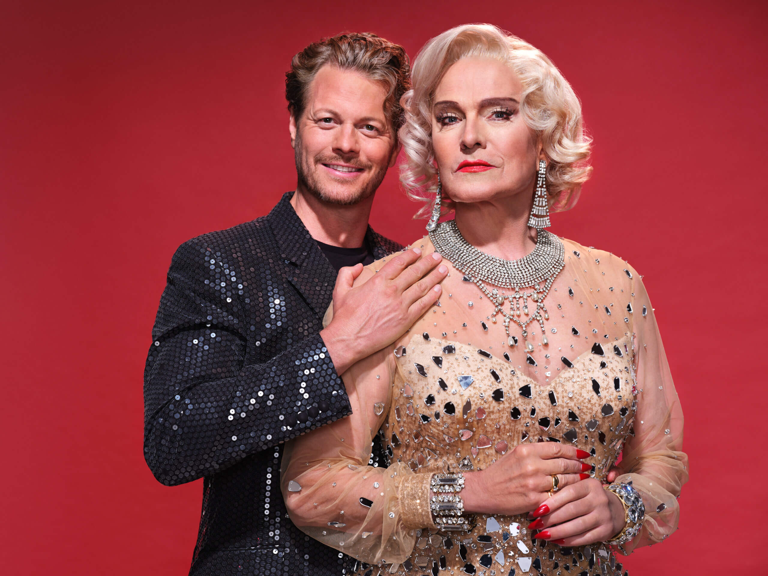 Zwei Personen in glamouröser Abendkleidung vor rotem Hintergrund, elegant posierend mit funkelndem Schmuck.