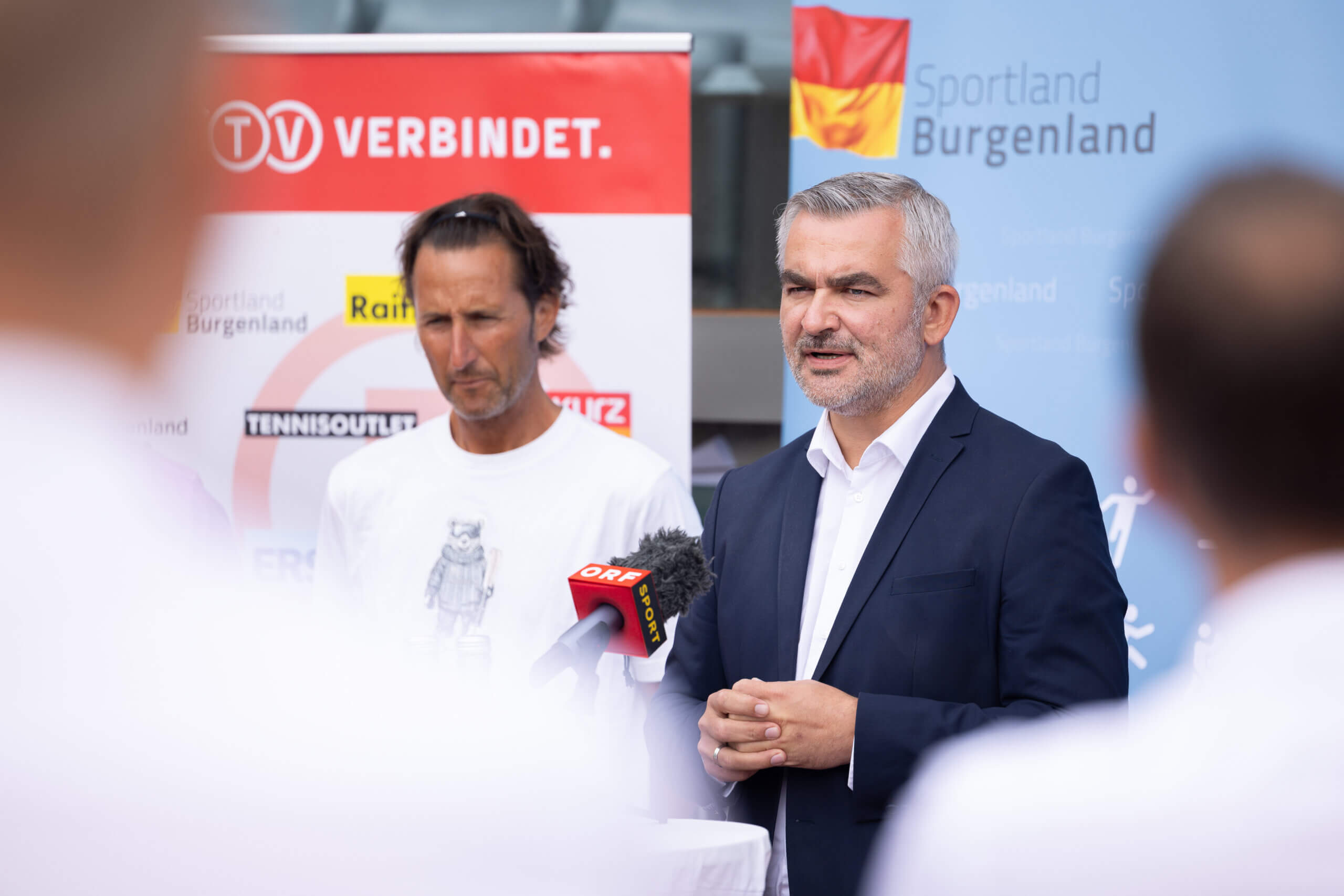 Ein Mann hält eine Rede vor einem Sportland Burgenland Poster. Medienvertreter hören aufmerksam zu.