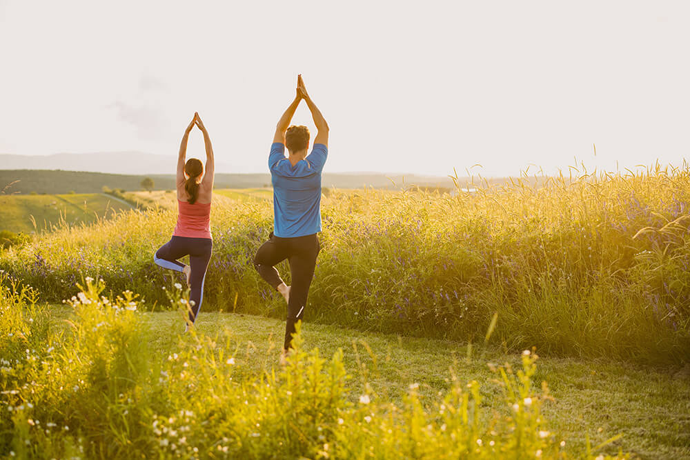 WEB_Yogaauszeit in Bad Tatzmannsdorf ©Bad Tatzmannsdorf Tourismus - K. Schrotter Photograph Zwei Personen praktizieren Yoga im Freien, umgeben von grünen Feldern und Sonnenlicht.