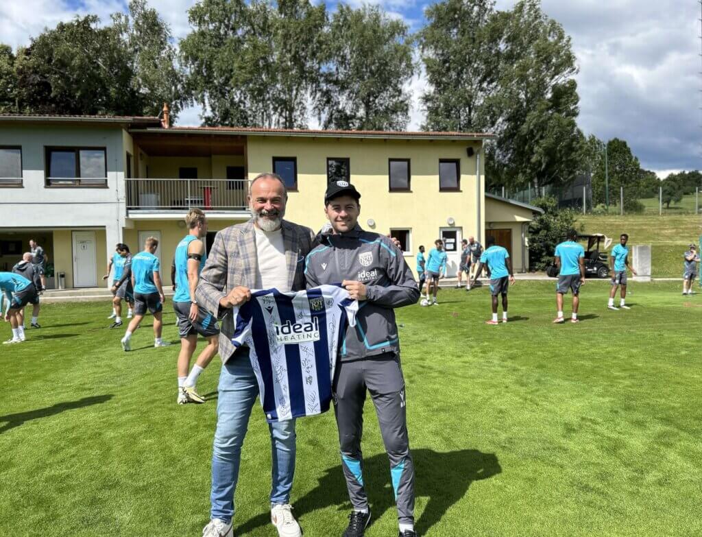 West Bromwich Albion Trainer Ryan Mason und AVITA GF Peter Prisching_©AVITA Resort_V.Baum Zwei Männer halten ein signiertes Fußballtrikot, während Fußballspieler im Hintergrund auf einem Trainingsplatz stehen.
