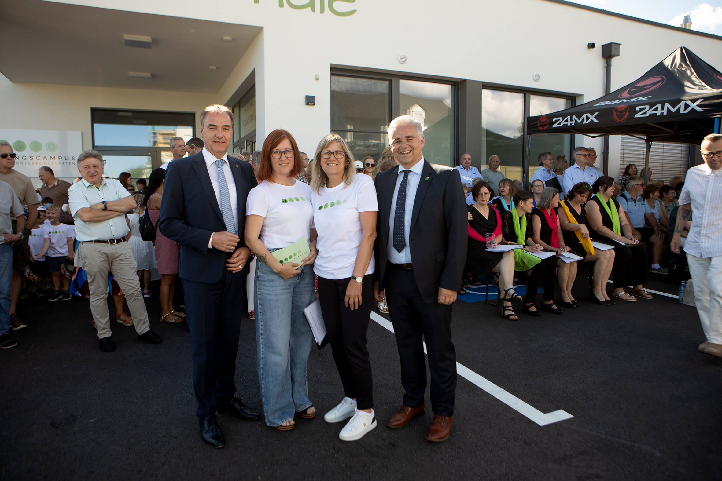 Gruppe von Menschen vor einem Gebäude bei einer Veranstaltung, einige tragen T-Shirts mit grünem Logo, andere Anzüge.