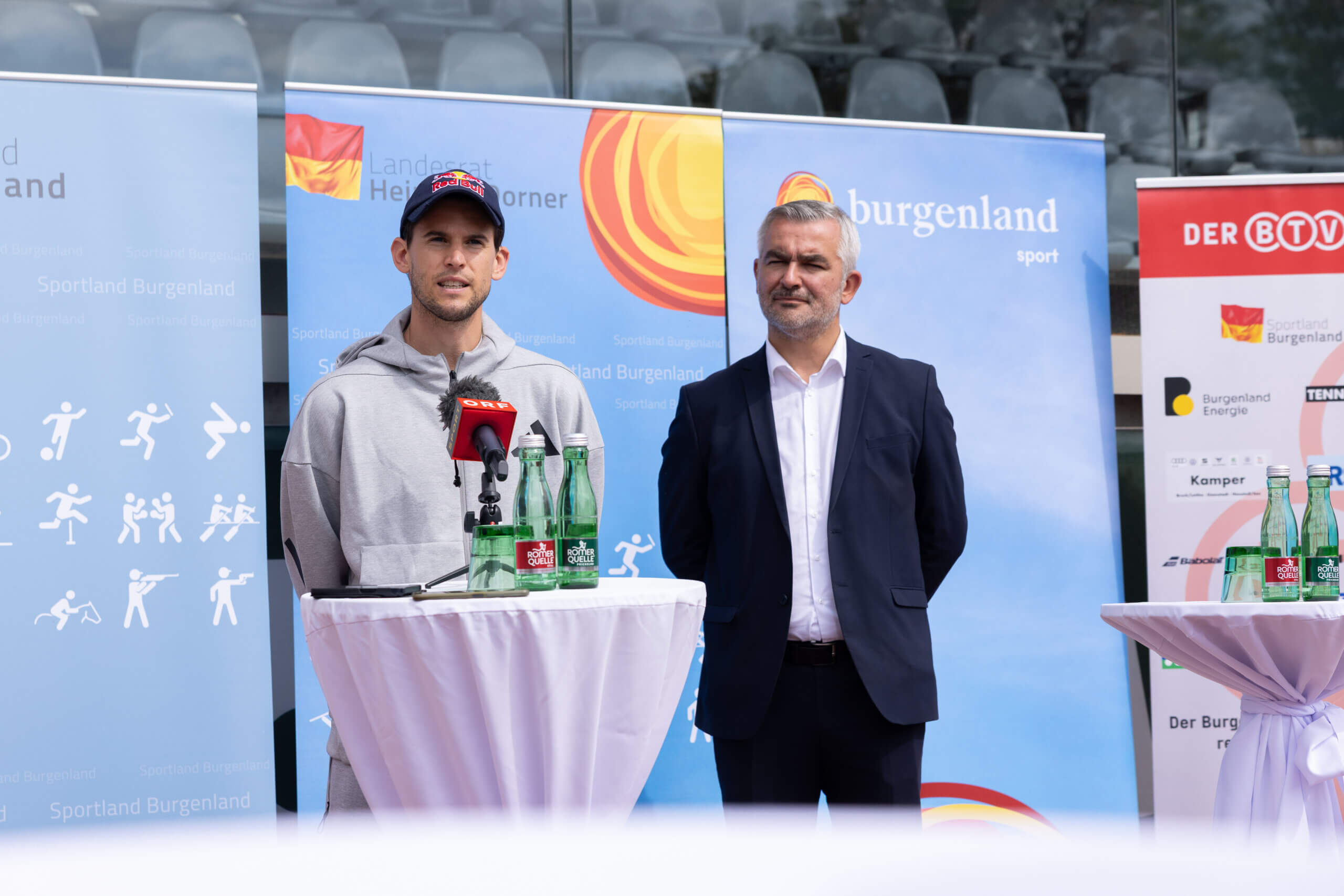 Sport-Pressekonferenz im Burgenland: Zwei Männer am Rednerpult mit Mikrofonen und Wasserflaschen, Sportlogos im Hintergrund.