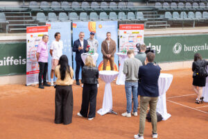 Pressekonferenz auf einem Tennisplatz im Burgenland mit mehreren Rednern und Zuhörern vor Sponsor-Bannern.