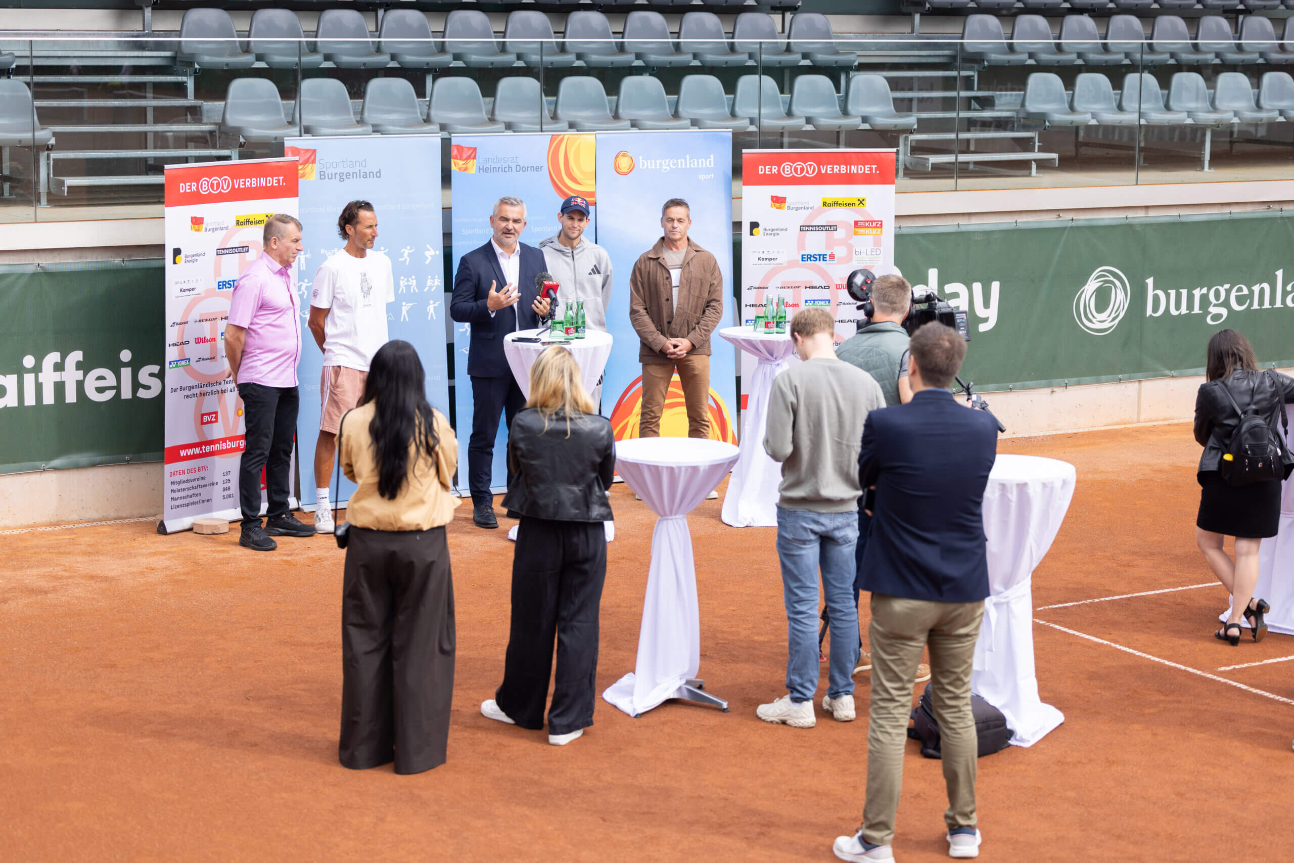 Pressekonferenz auf einem Tennisplatz im Burgenland mit mehreren Rednern und Zuhörern vor Sponsor-Bannern.