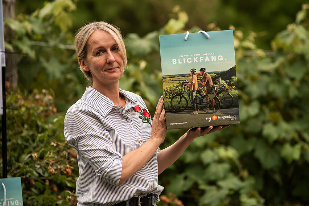 Frau hält eine Burgenland-Werbetasche im Freien, mit Radfahrermotiv und Blickfang-Aufschrift, umgeben von grüner Natur.