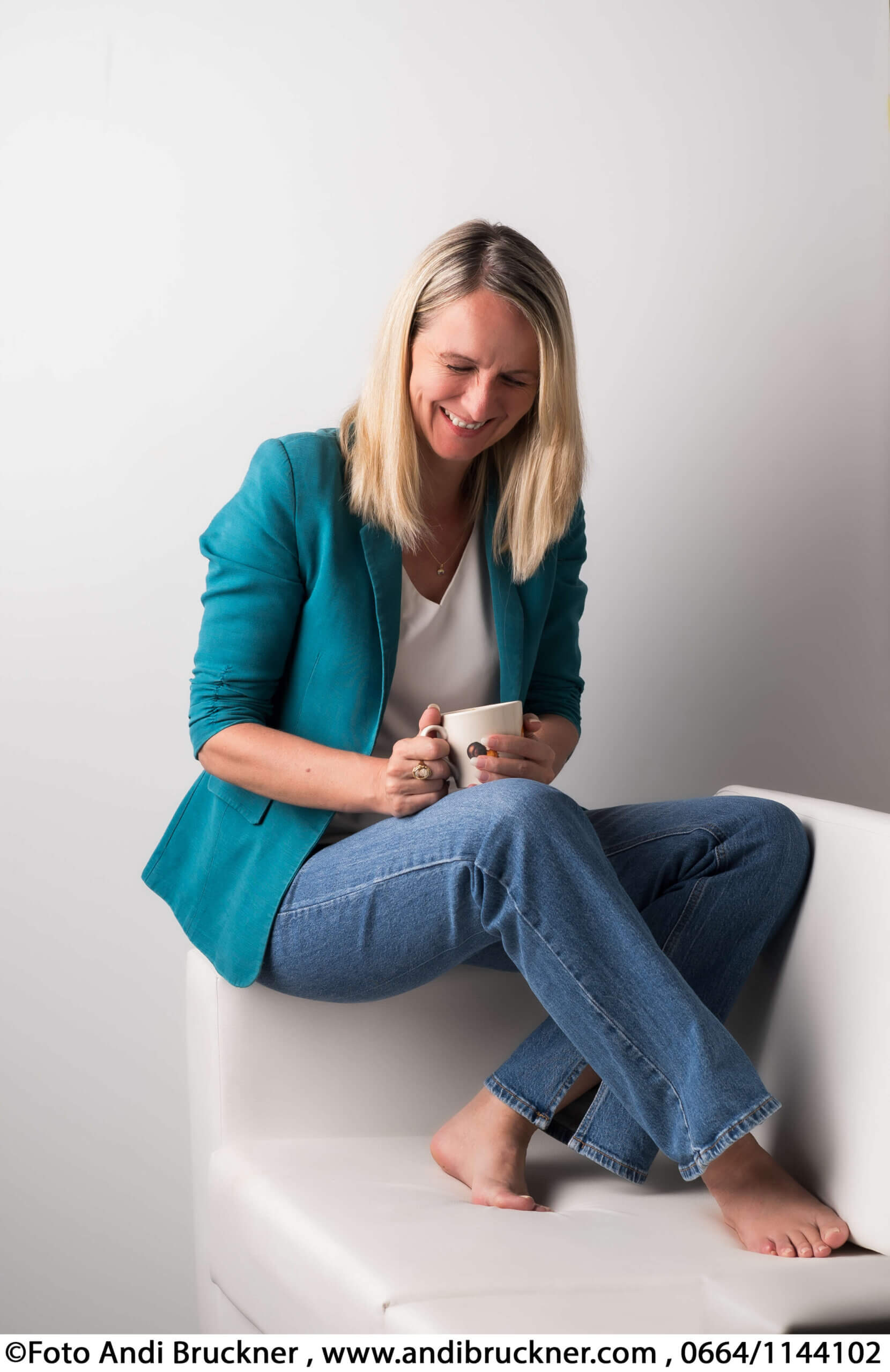 Lächelnde Frau in blauer Jacke, entspannt mit Kaffeetasse auf weißem Sofa sitzend.