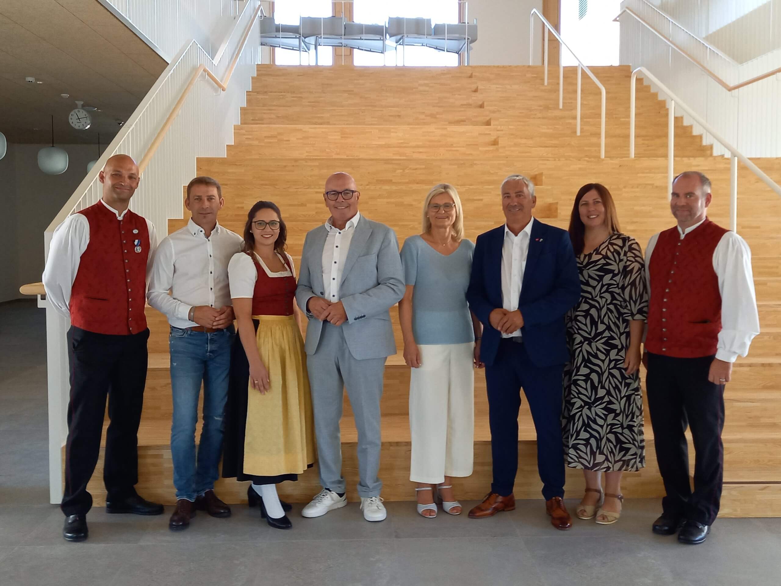 Gruppe von acht Menschen posiert in traditioneller und Business-Kleidung vor einer modernen Treppe in einem hellen Raum.