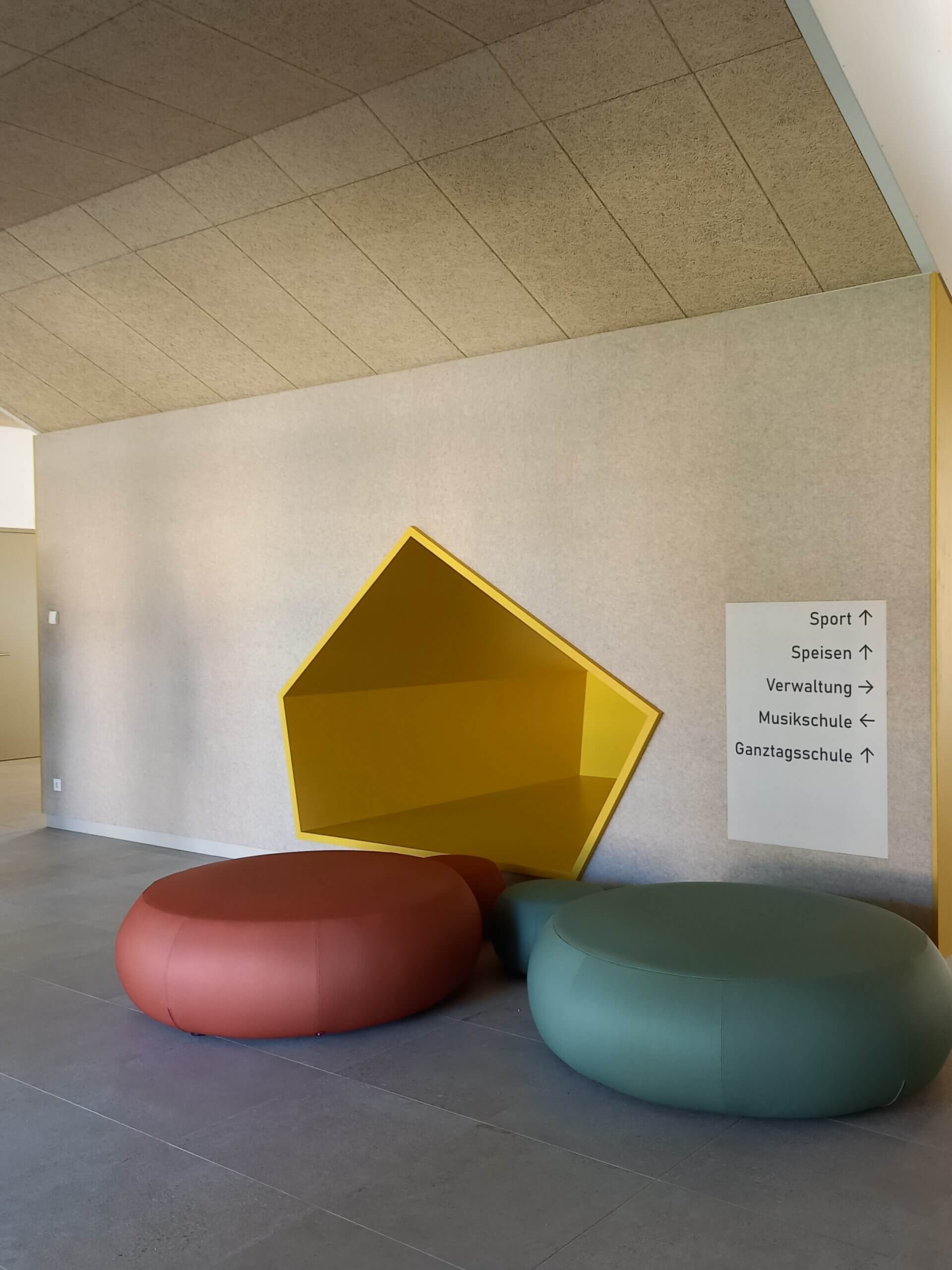 Moderne Sitzecke in der Schule mit bunten Ottomane, gelbem Wandregal und Wegweiser zu verschiedenen Bereichen.