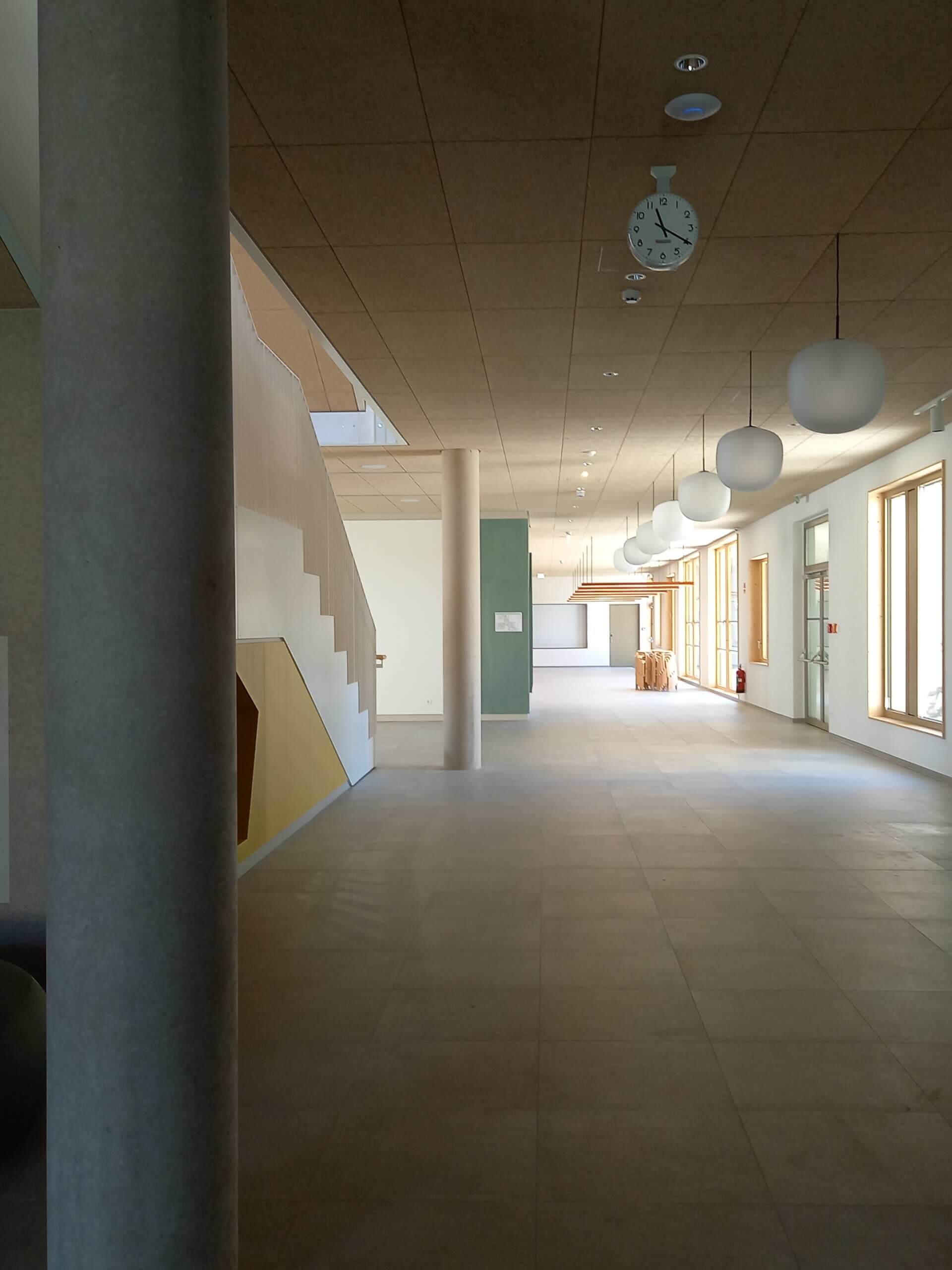 Moderner Büroflur mit Treppe, Hängelampen und großen Fenstern, die natürliches Licht hereinlassen.