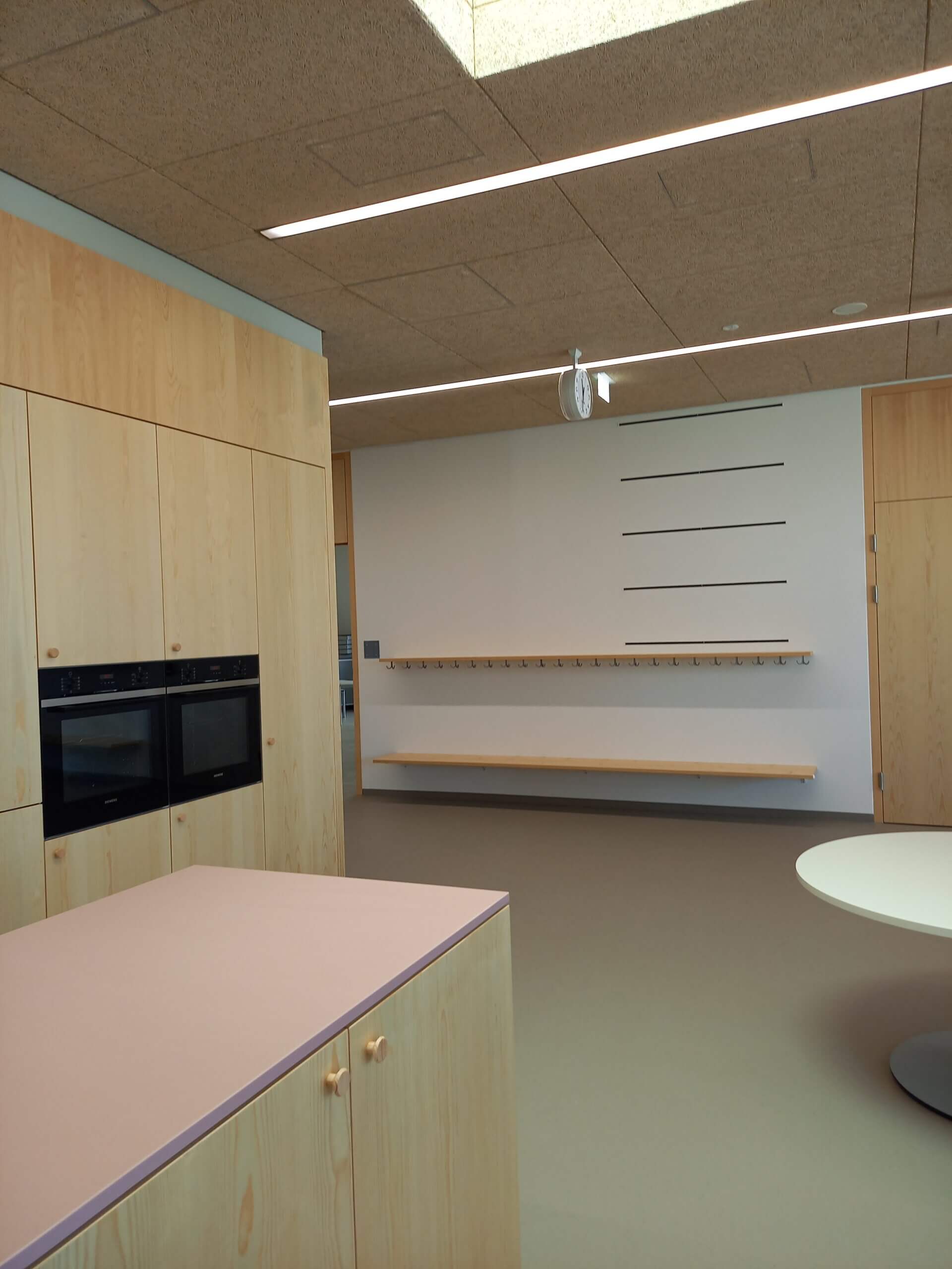 Moderne Küche mit Holzmöbeln, Ofen und Tisch, beleuchtet von Deckenleuchten, minimalistisch und funktionell designt.