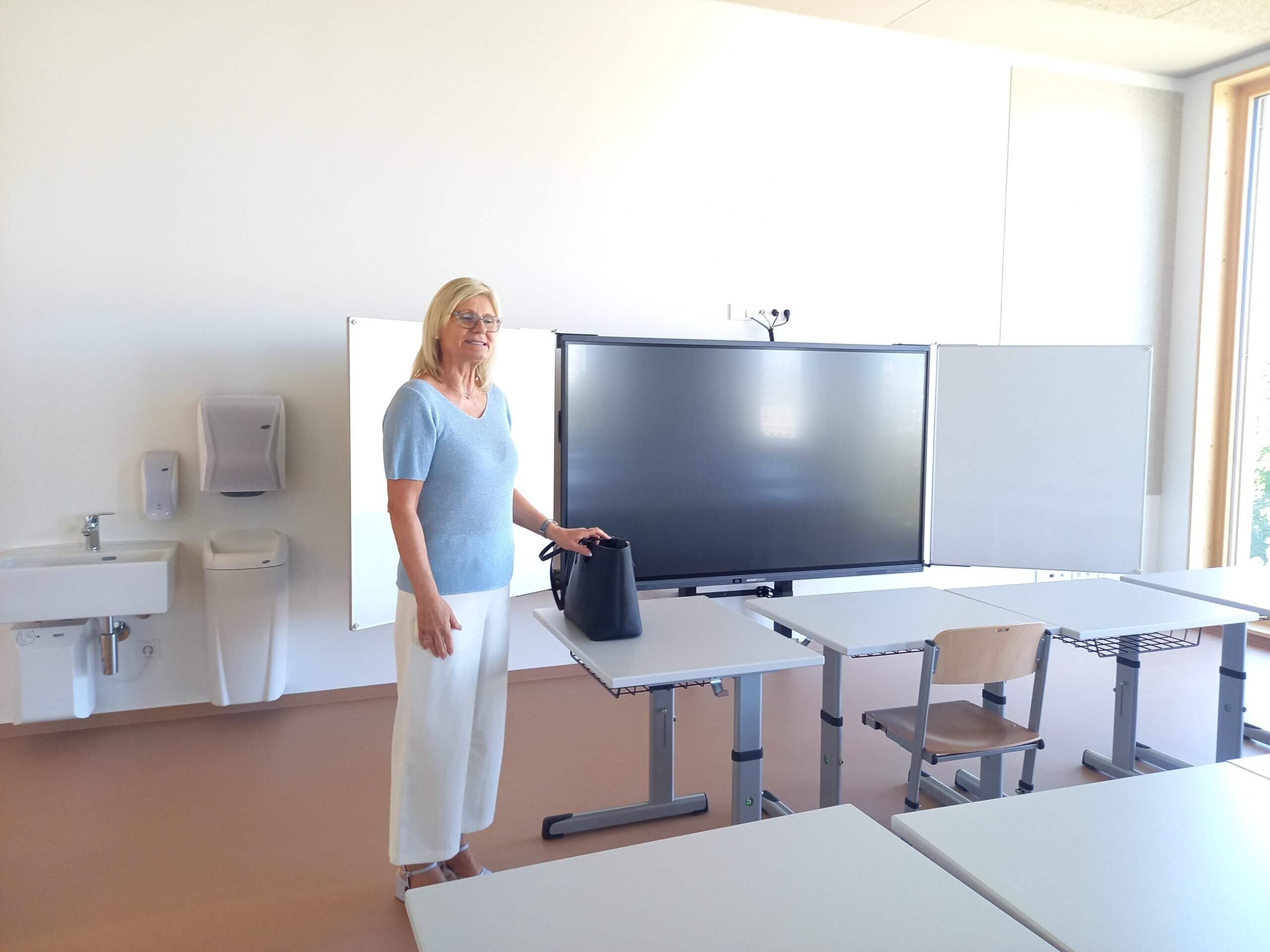 Frau in einem modernen Klassenzimmer mit Smartboard und Schultischen, bereit für den Unterricht.