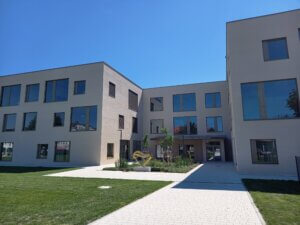 Moderne Schulgebäude-Architektur bei Sonnenschein, mit großzügigen Glasfenstern und gepflegtem Gartenbereich.