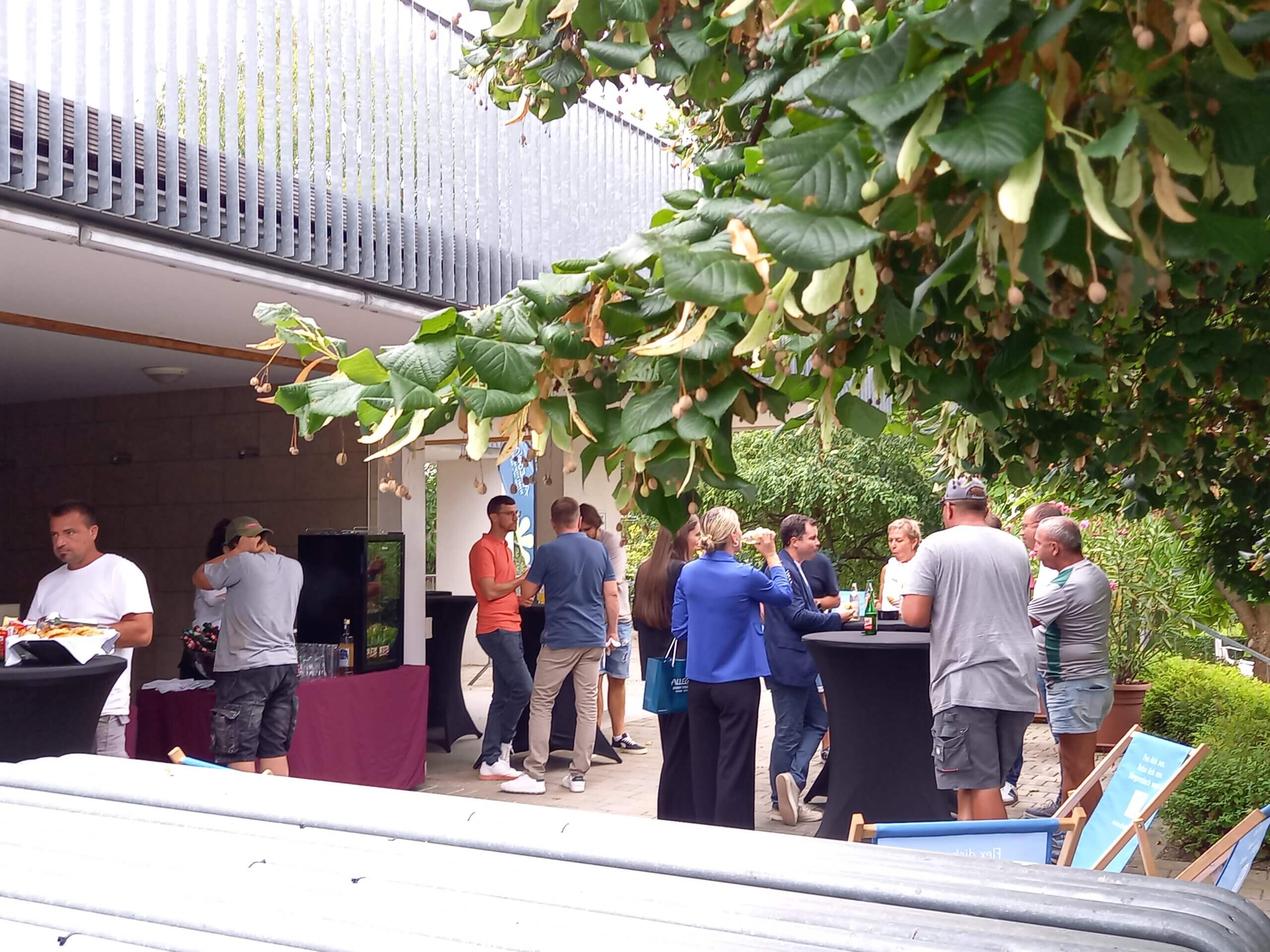 Menschen treffen sich im Freien für Networking, umgeben von Bäumen und Tischen mit Getränken.