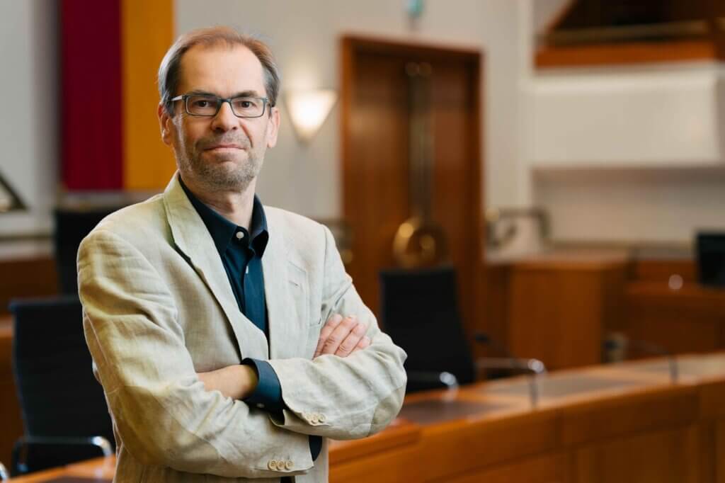 Mann in beigeem Sakko steht mit verschränkten Armen in einem Konferenzraum und schaut in die Kamera. Foto zum Thema Klimagesetz