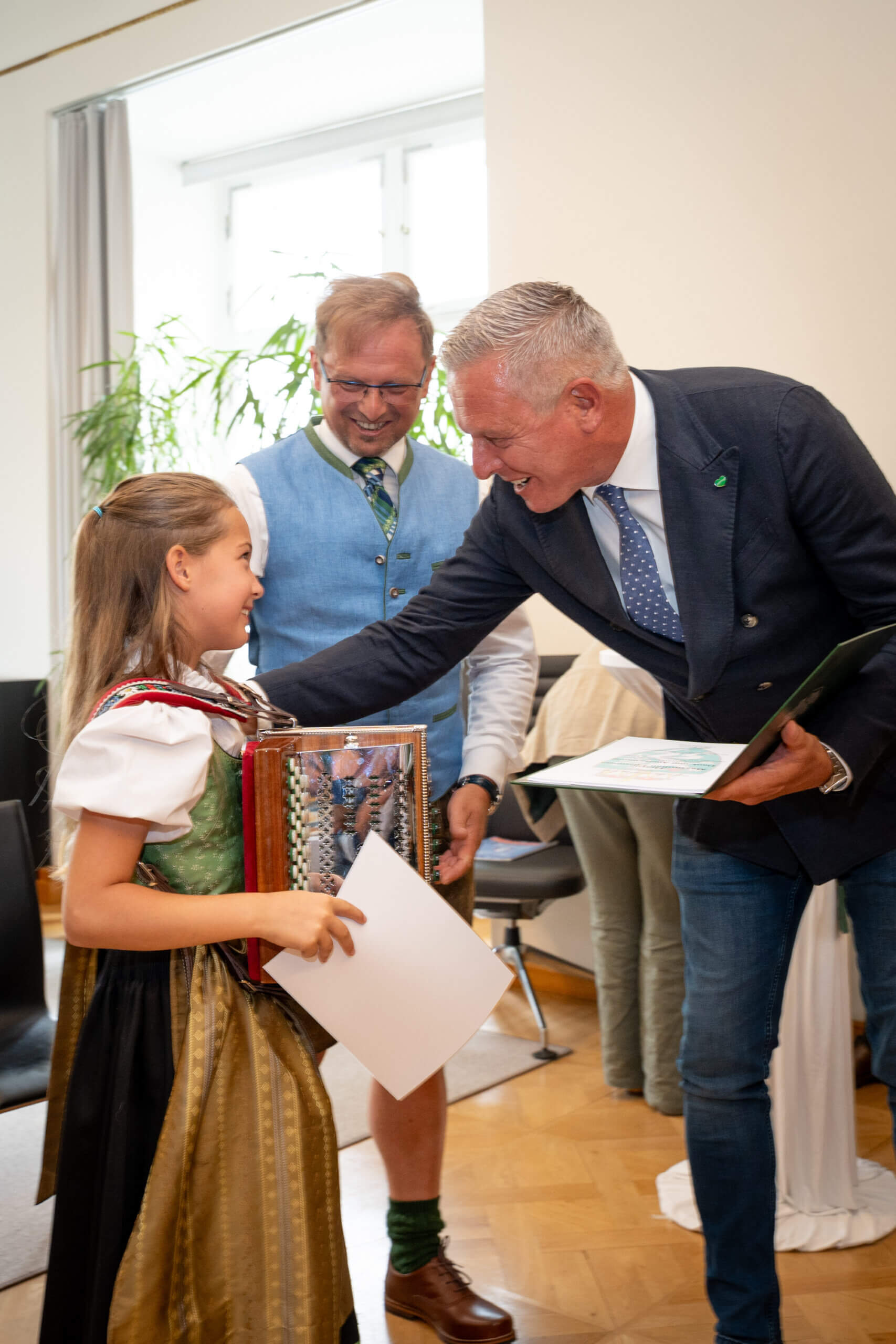 Mädchen im Dirndl erhält Auszeichnung von zwei Männern bei einer Feier, lächelnd und mit Akkordeon in der Hand.