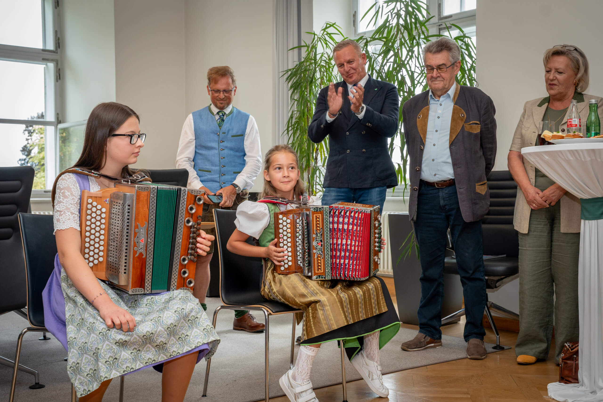 Mädchen spielen Akkordeon in traditioneller Kleidung, während Erwachsene applaudieren in einem Festsaal.