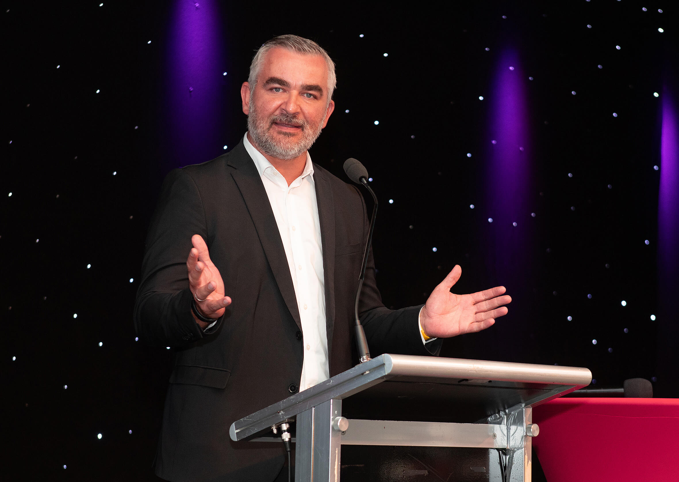 Redner am Podium unter lila Licht, gestikulierend vor einer schwarzen Bühne mit Sternenhintergrund.