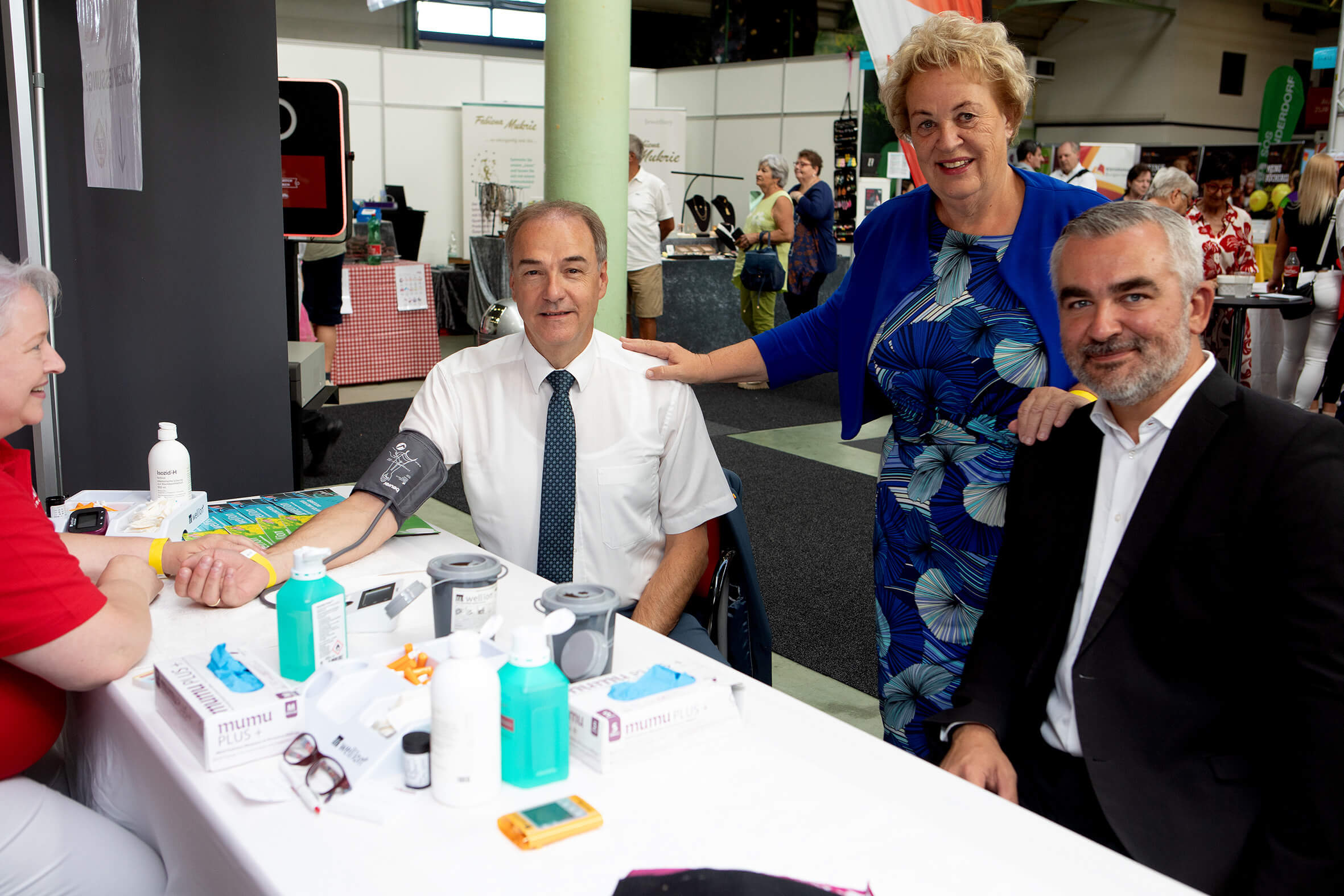 Personen bei einer Gesundheitsexpo, Blutdruckmessung am Stand, freundliches Gespräch und Beratung.