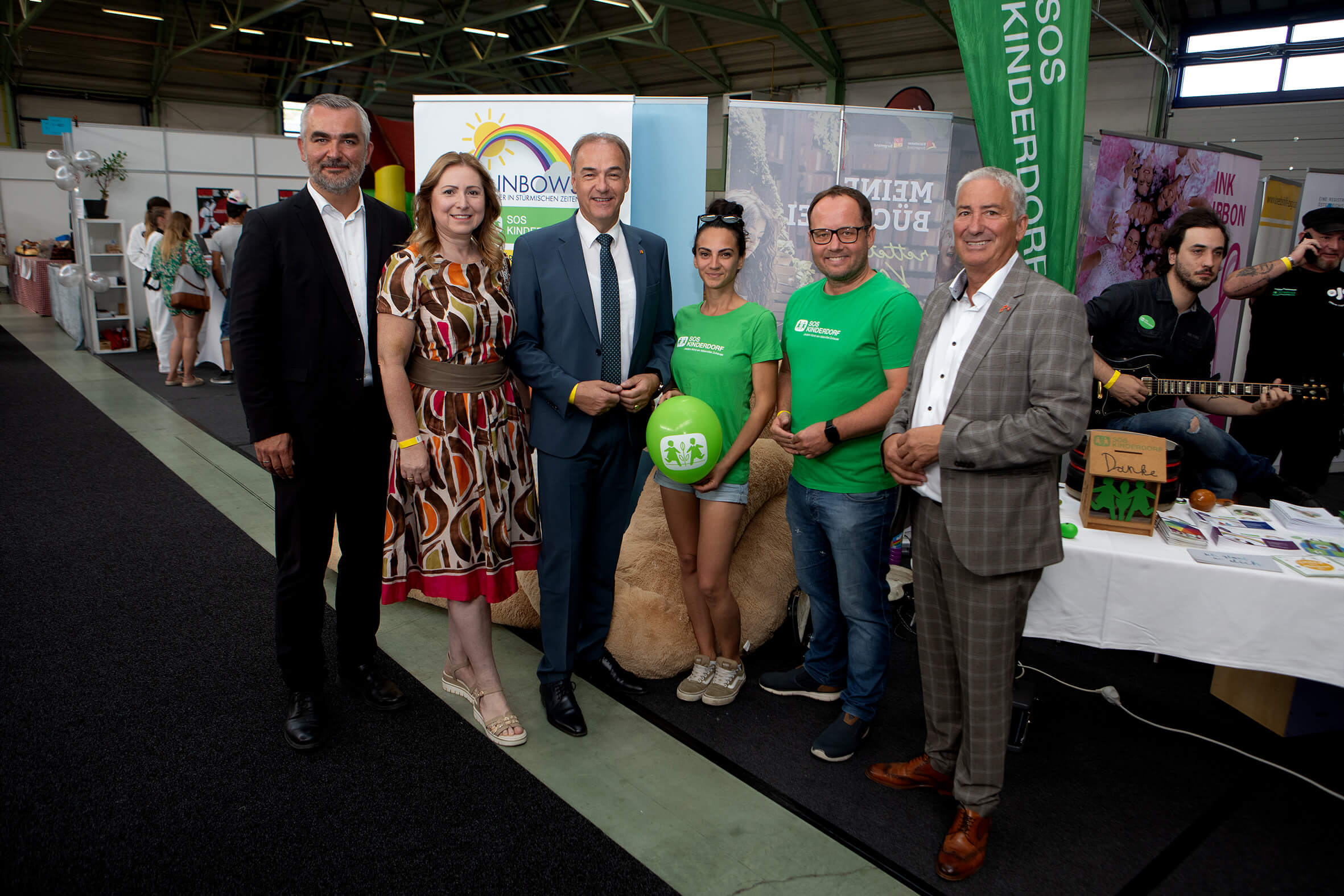 Gruppe von Menschen posiert bei einer Messeveranstaltung, SOS-Kinderdorf-Stand im Hintergrund, fröhliche Atmosphäre.