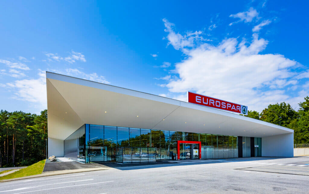 Moderne Eurospar-Filiale mit Glasfassade unter blauem Himmel, umgeben von grünen Bäumen.