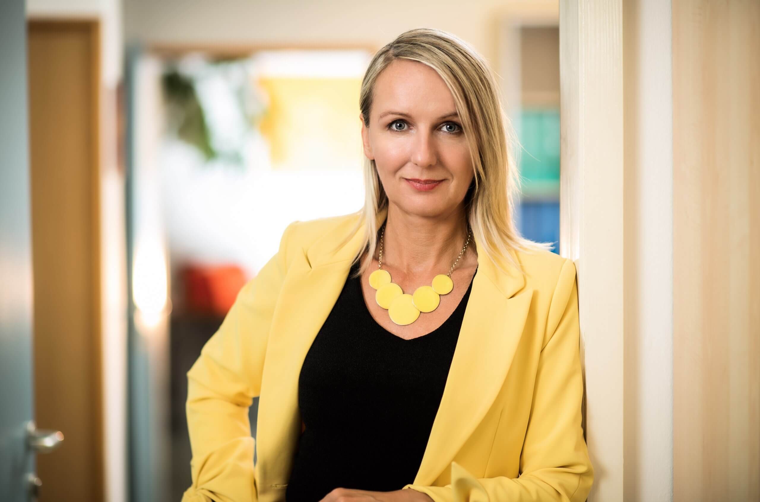 Frau im gelben Blazer und modernem Schmuck lehnt an einer Bürotür, lächelnd und selbstbewusst im Geschäftsumfeld.