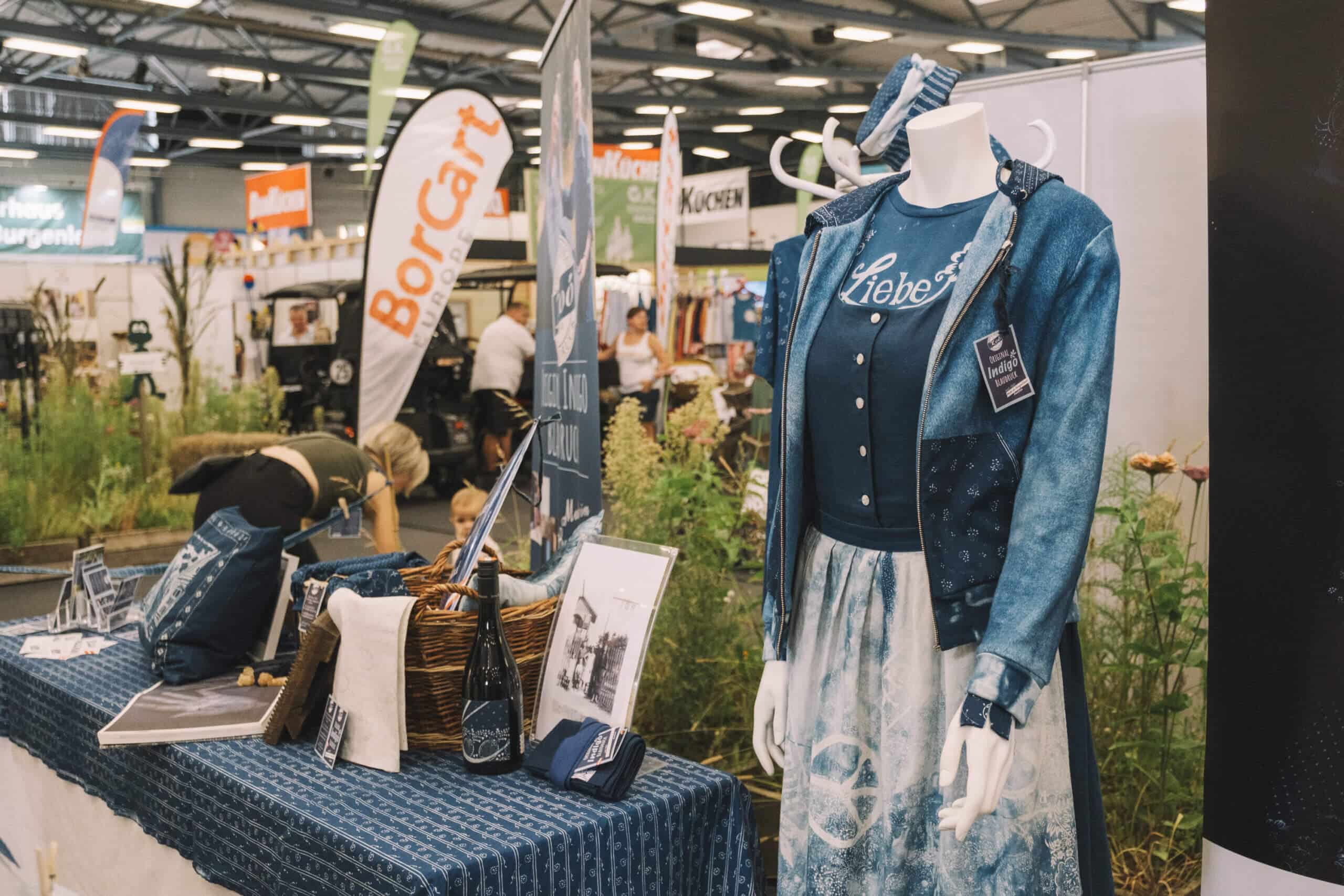 Puppe in traditionellem Denim-Outfit auf Messe, umgeben von Deko und Werbematerialien.