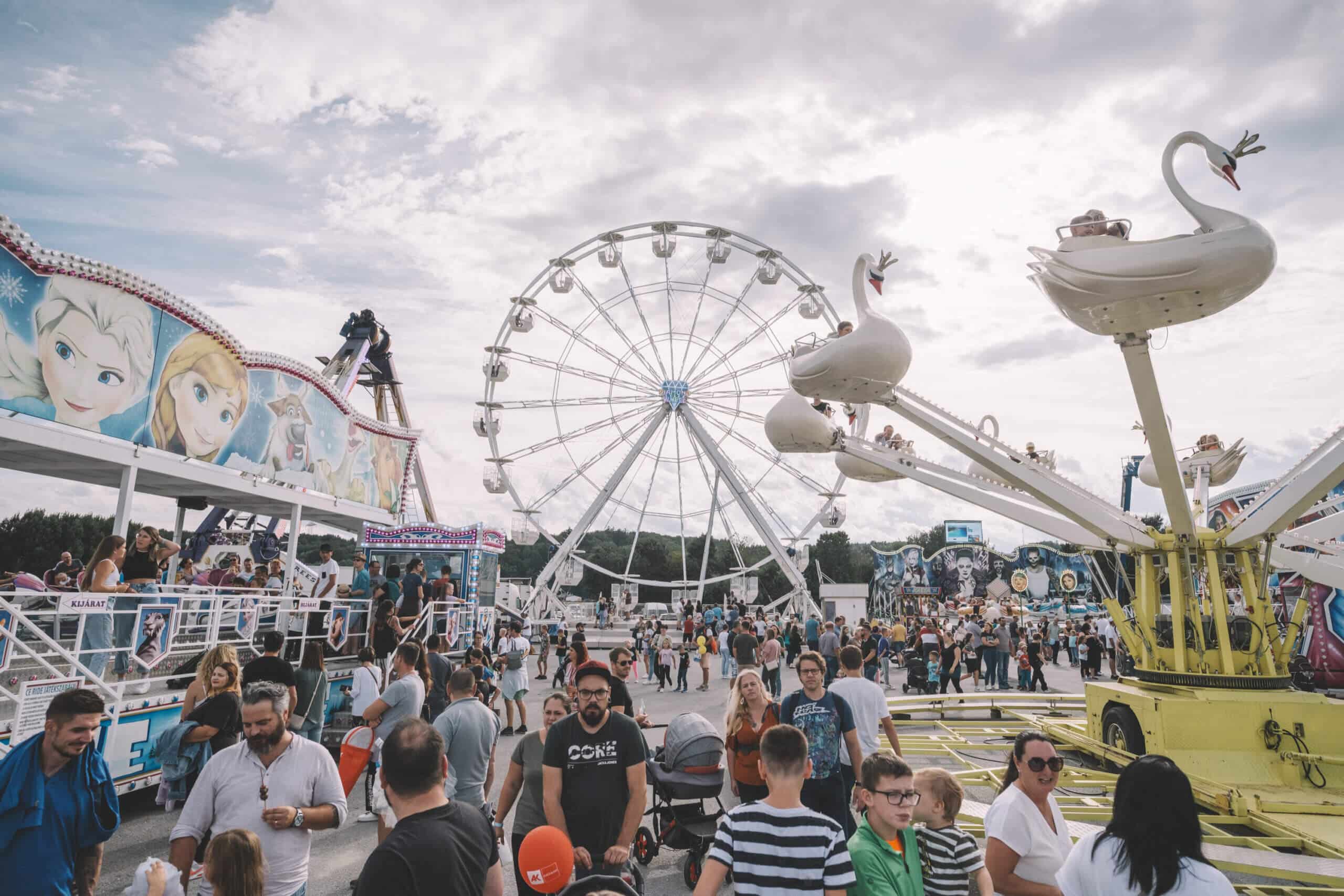 Fröhliche Menschenmenge auf Jahrmarkt mit Riesenrad und Schwanenkarussell unter bewölktem Himmel.