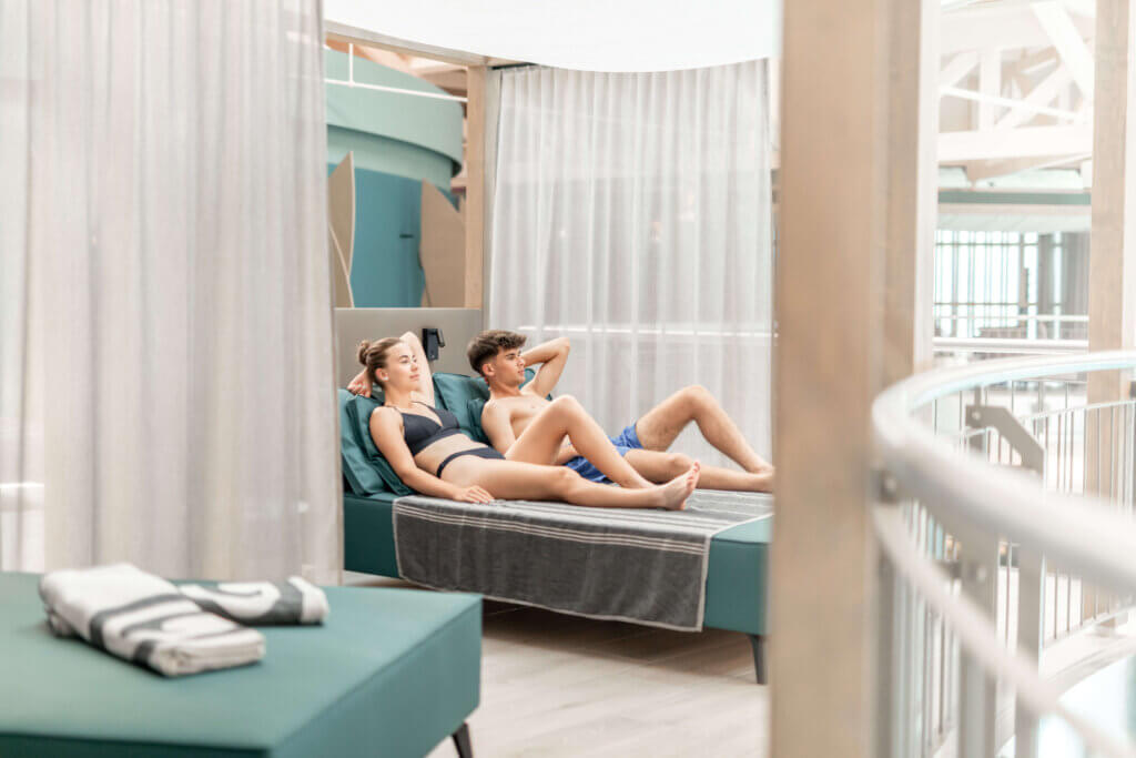 Deluxe Loungen_©AVITA Resort Paar entspannt auf Liege im Wellnessbereich, umgeben von hellen Vorhängen und moderner Architektur.