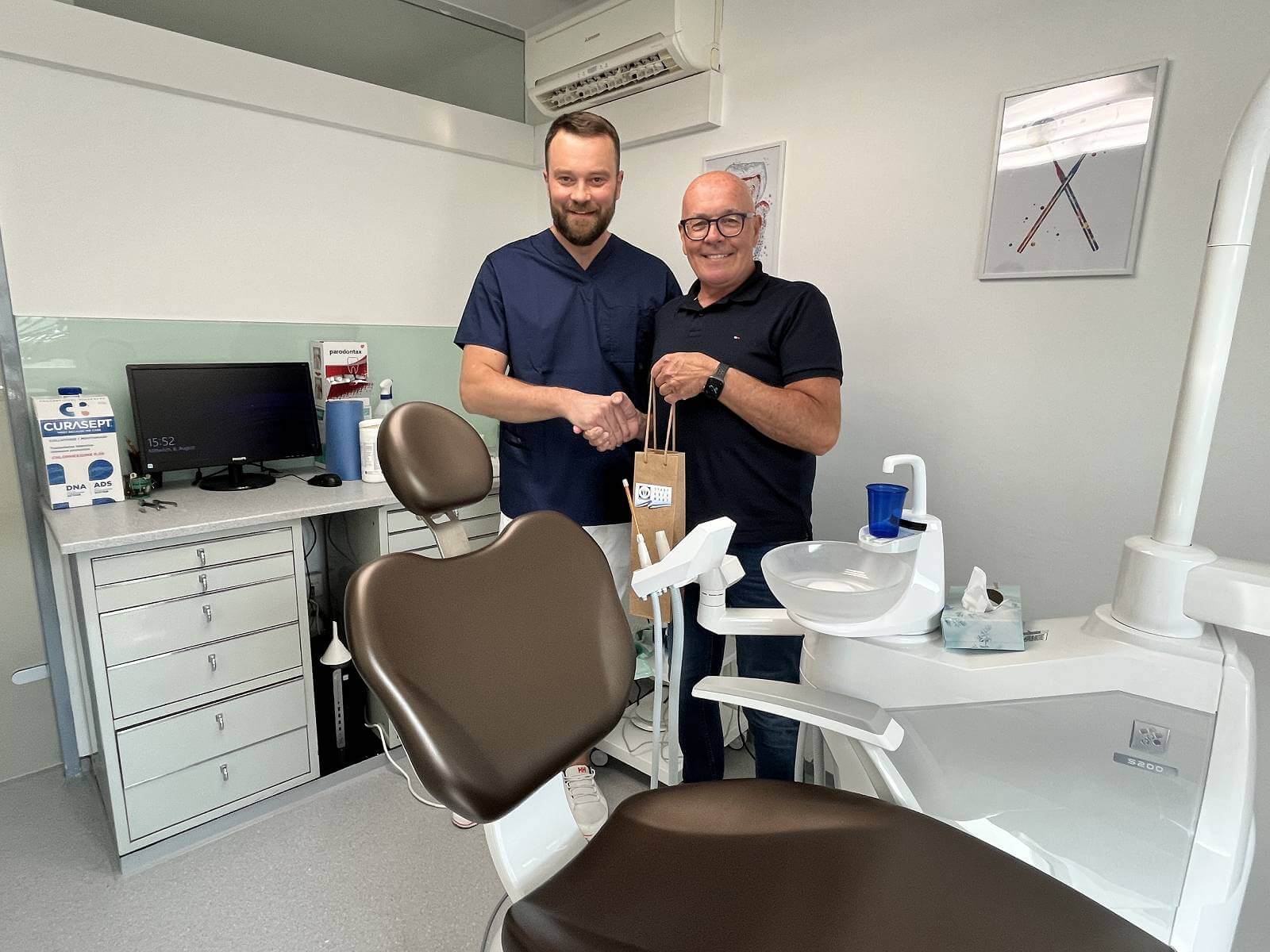 Zahnarzt und Patient schütteln Hände in moderner Zahnarztpraxis mit Behandlungsstuhl im Vordergrund.