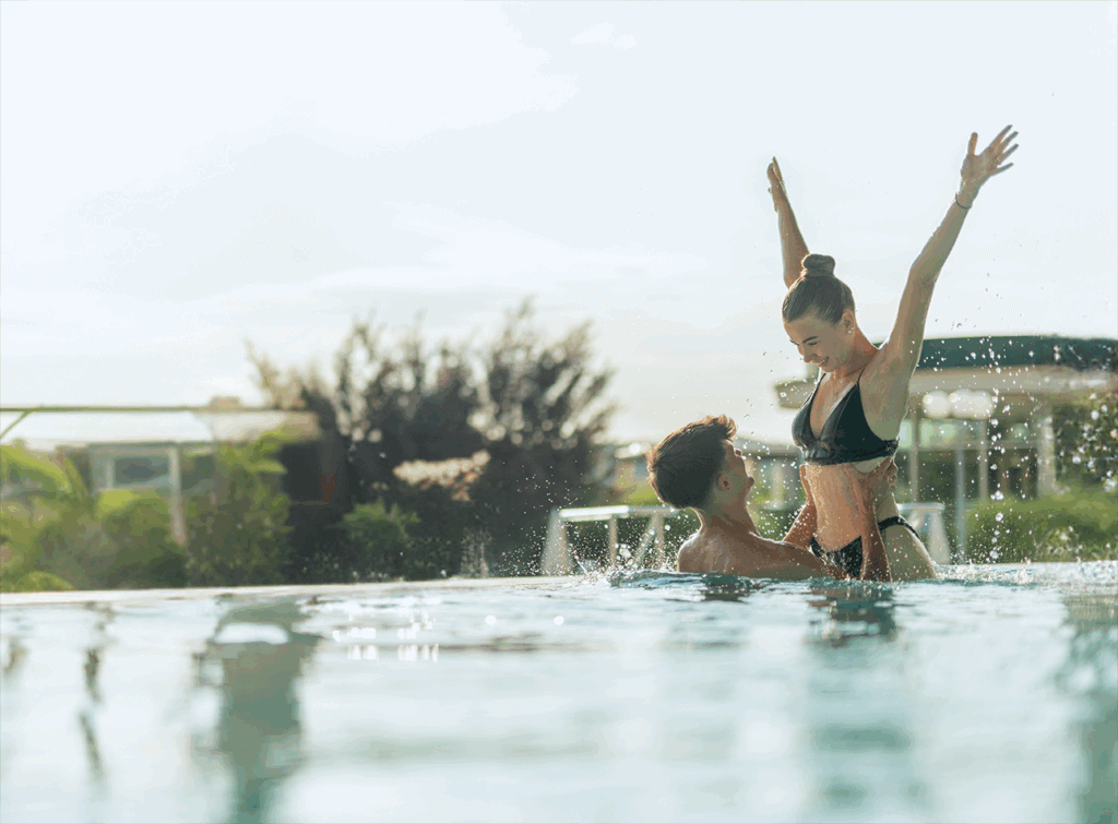 Eine Therme zum Verlieben_©AVITA Resort Paar genießt Spaß im Pool an einem sonnigen Tag, spritzt Wasser und lacht, Sommerfreude im Freien.